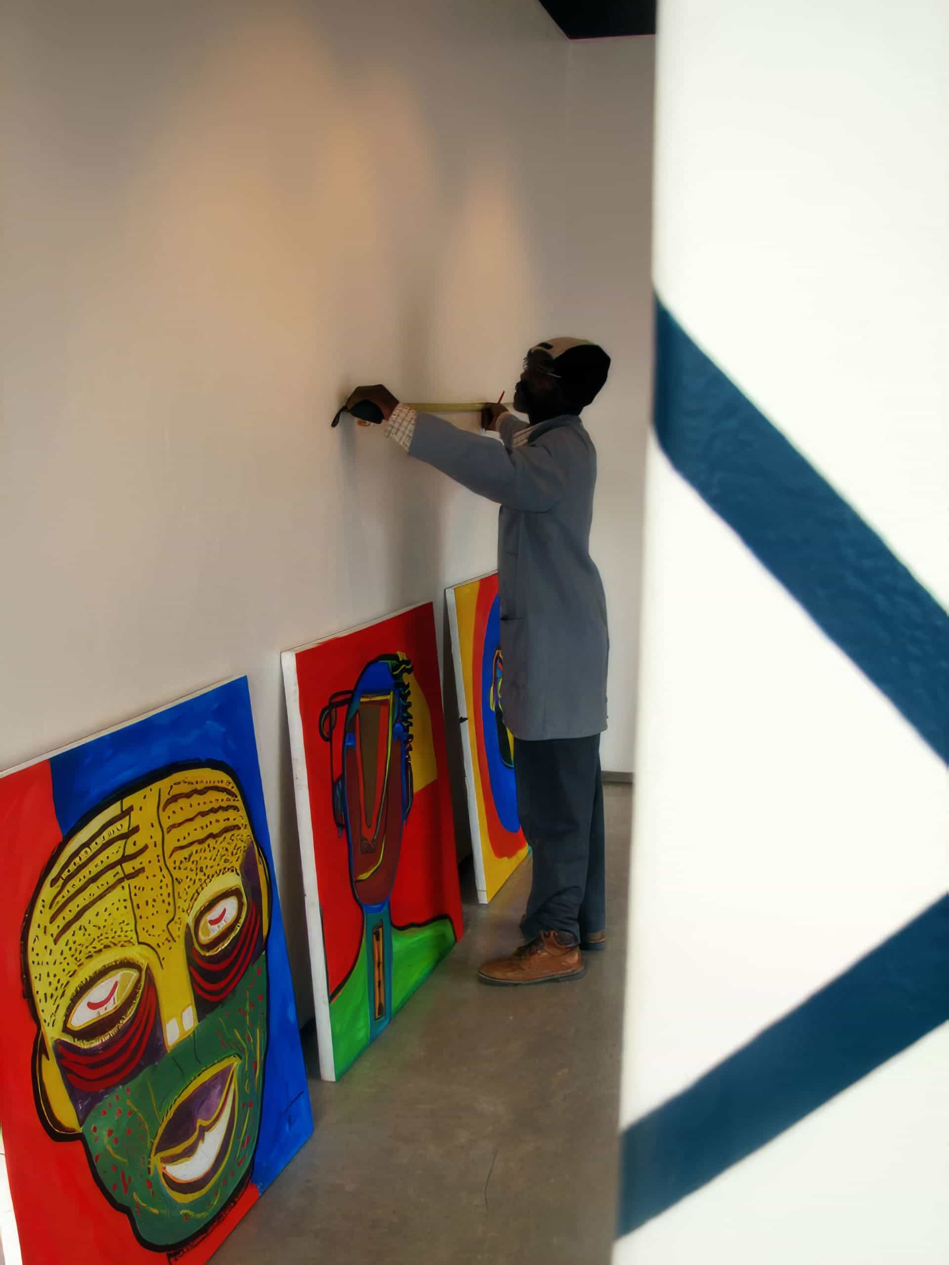 A person in a grey jacket hangs artwork on a white gallery wall. Three colourful abstract paintings lean against the wall below, depicting stylised faces in bold reds, blues, greens, and yellows. A white pillar with a blue stripe is in the foreground.