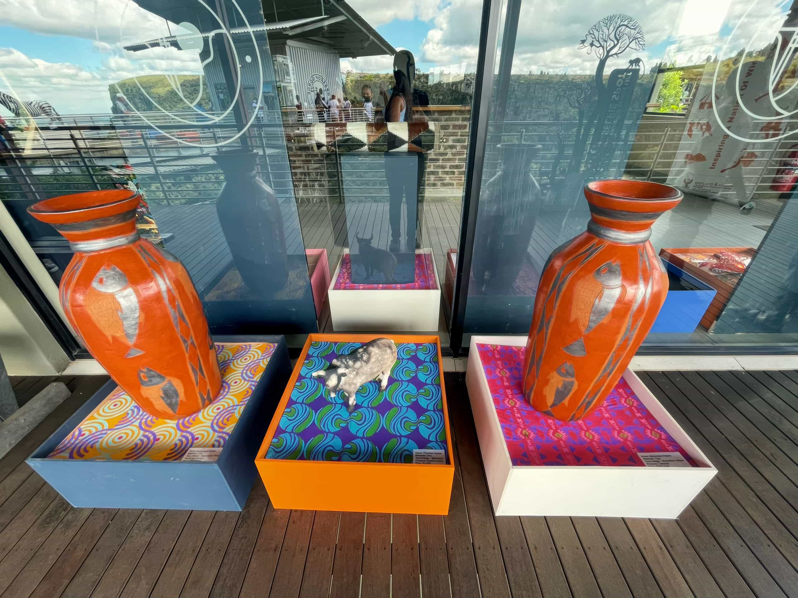 Two large orange vases with fish designs stand on colourful patterned platforms. Between them, a grey and white cat lies curled up on a pink and blue plinth. A reflection of a person taking the photo appears in the glass behind the display.