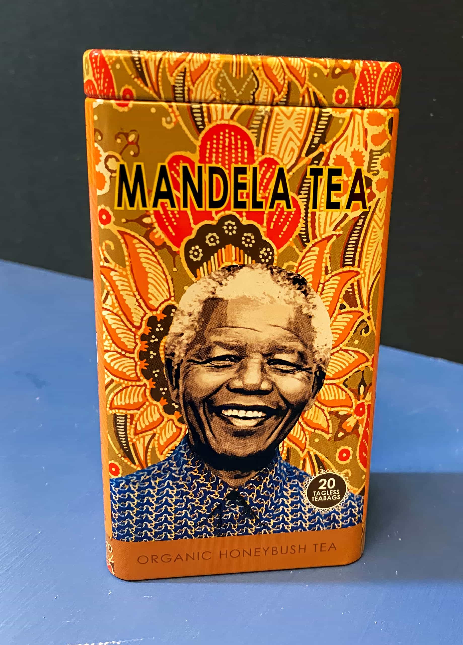 A decorative tin of Mandela Tea features a smiling portrait of Nelson Mandela. The tin is orange with intricate, colourful patterns and text reading 