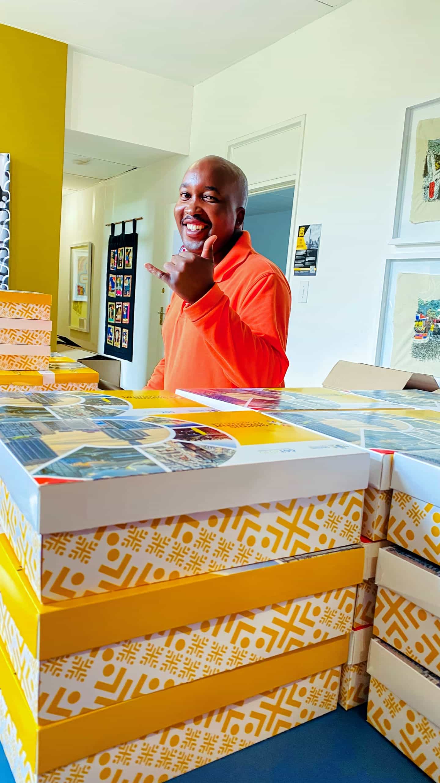 A smiling person in an orange shirt stands behind a table stacked with yellow-and-white patterned boxes. The person holds up a hand in a “hang loose” gesture, with colourful artwork and a yellow accent wall visible in the background.