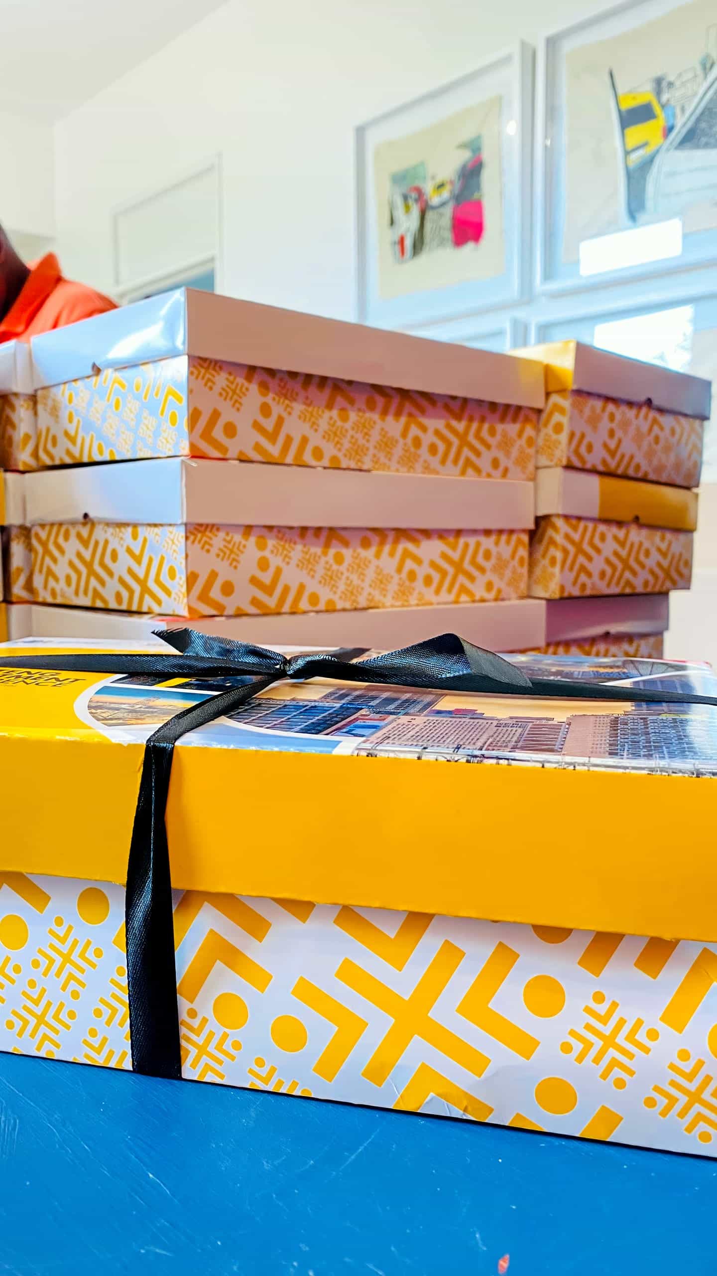 A stack of decorative yellow and white patterned boxes rests on a table, with one box in the foreground tied with a black ribbon. The background shows framed artwork on a white wall and part of a person out of focus.