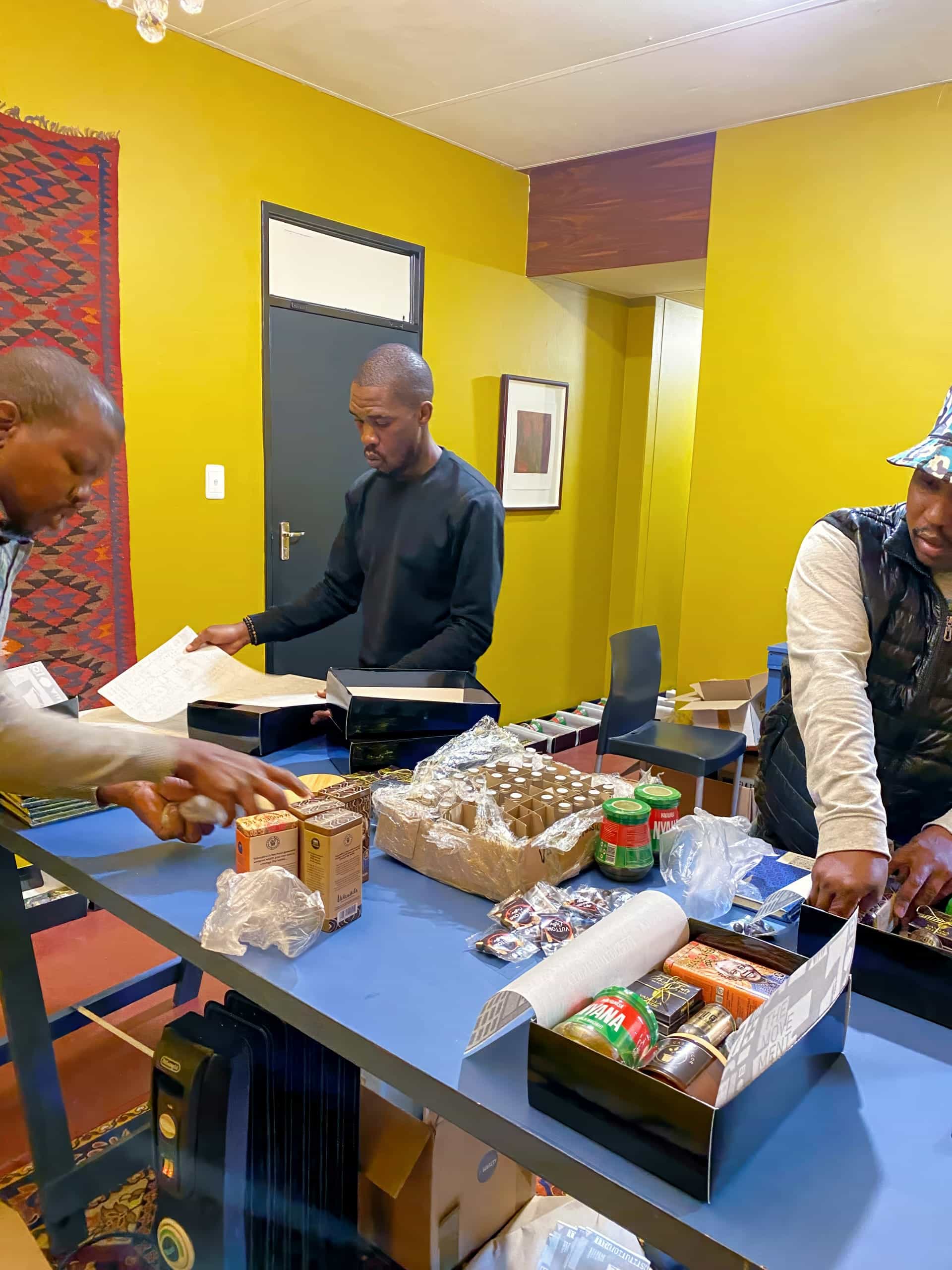 Three men stand at a blue table in a yellow-walled room, organising various food items, tins, jars, and papers. Shelves hold more supplies. The scene suggests teamwork and preparation, possibly for food distribution or community aid.