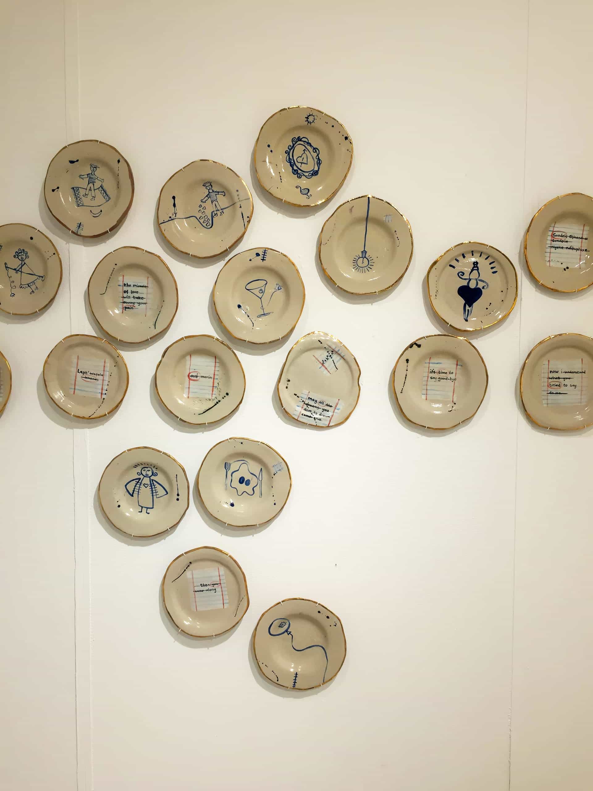 Twenty ceramic plates with drawings in blue are mounted on a white wall. The drawings depict simple, playful human or animal figures and objects. Some plates have handwritten text on lined paper, whilst others only have drawings. Plates are arranged loosely in a group.