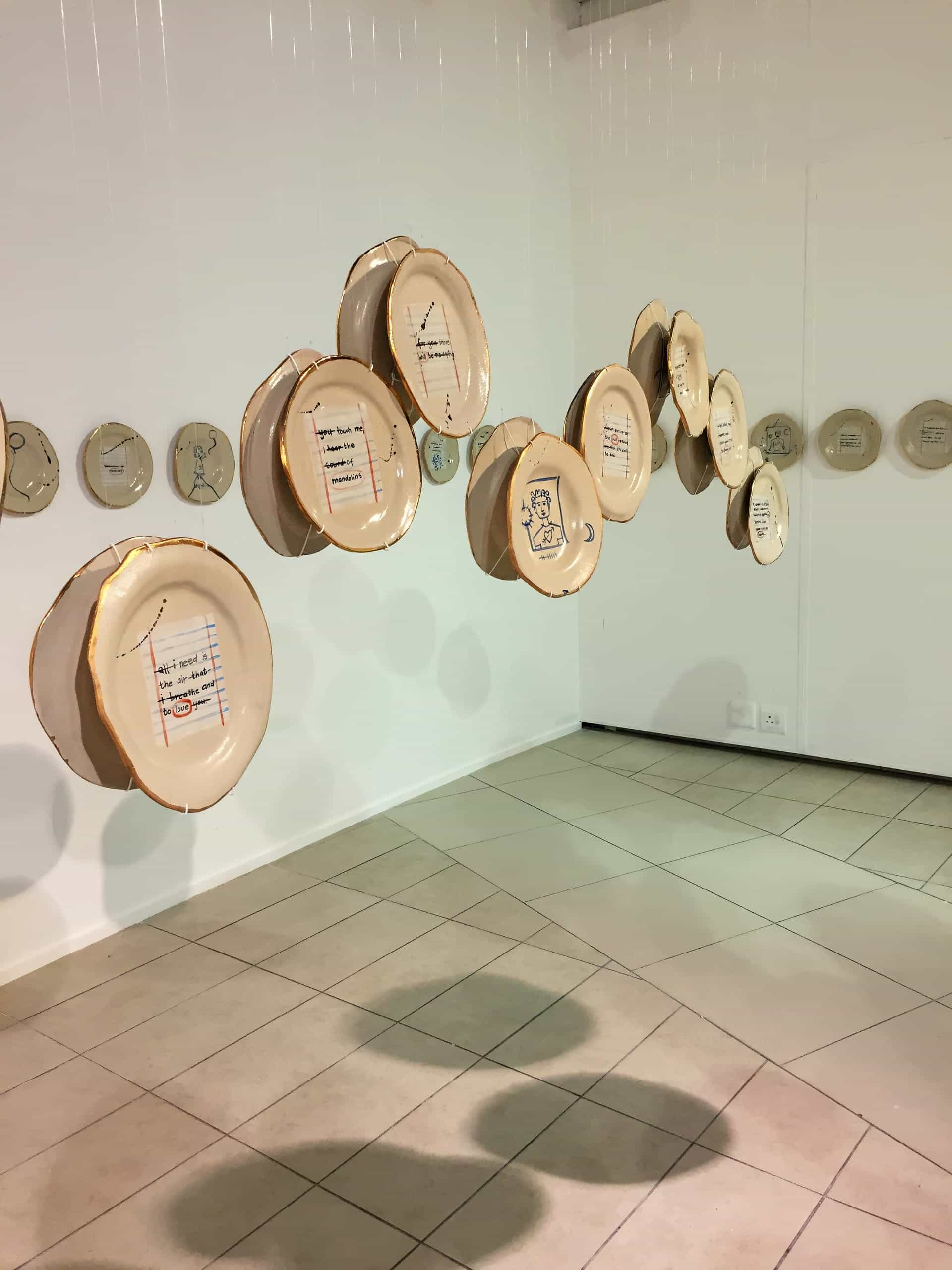 Several large, round ceramic plates with artistic drawings and writing are suspended in mid-air in a white gallery space. More plates with similar designs are mounted in a horizontal line along the wall in the background. The floor is tiled.