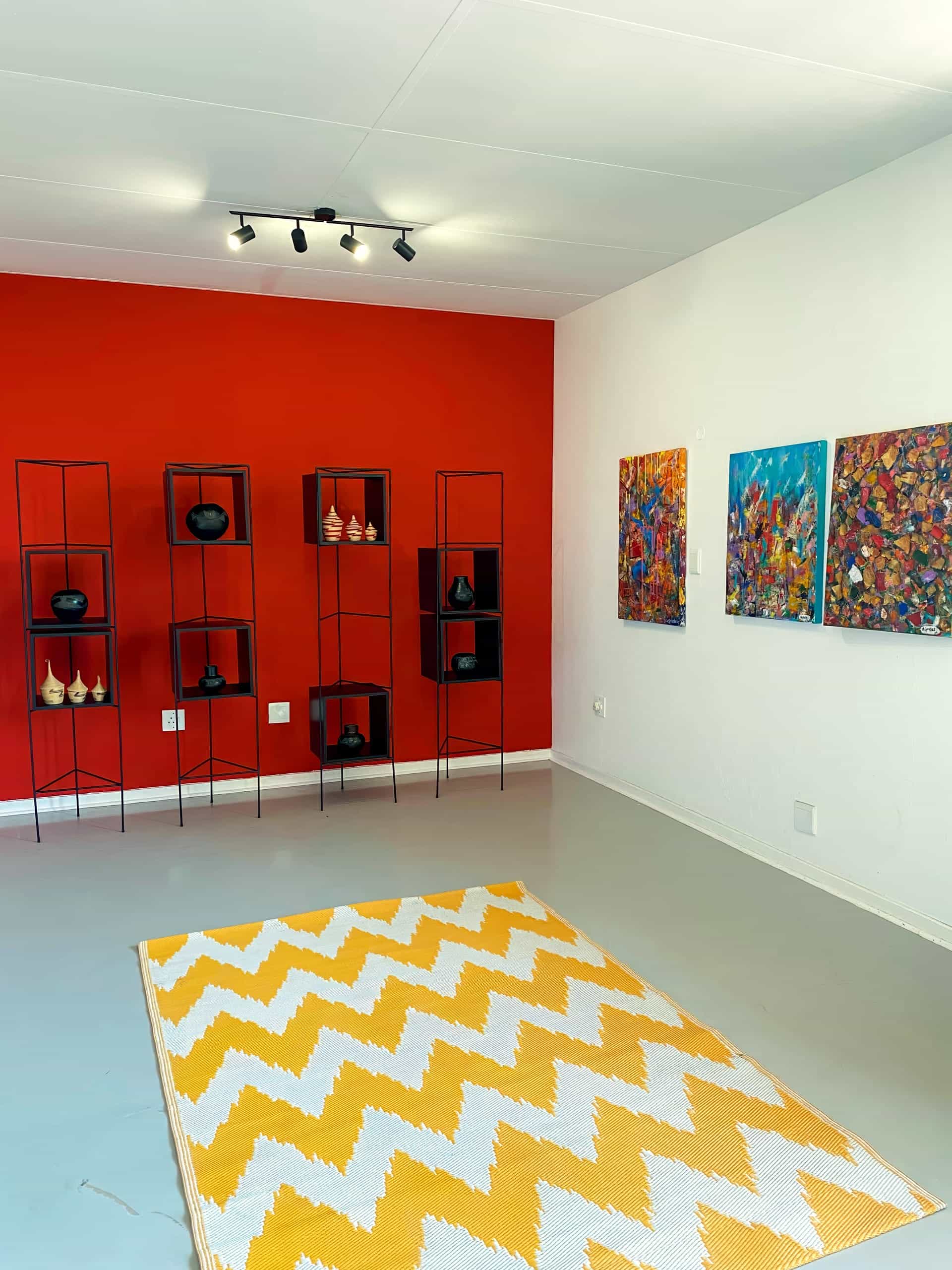 A modern art gallery with a red accent wall displaying pottery on black shelves, three colourful abstract paintings on a white wall, grey flooring, and a yellow and white chevron rug in the centre. Ceiling track lights illuminate the space.