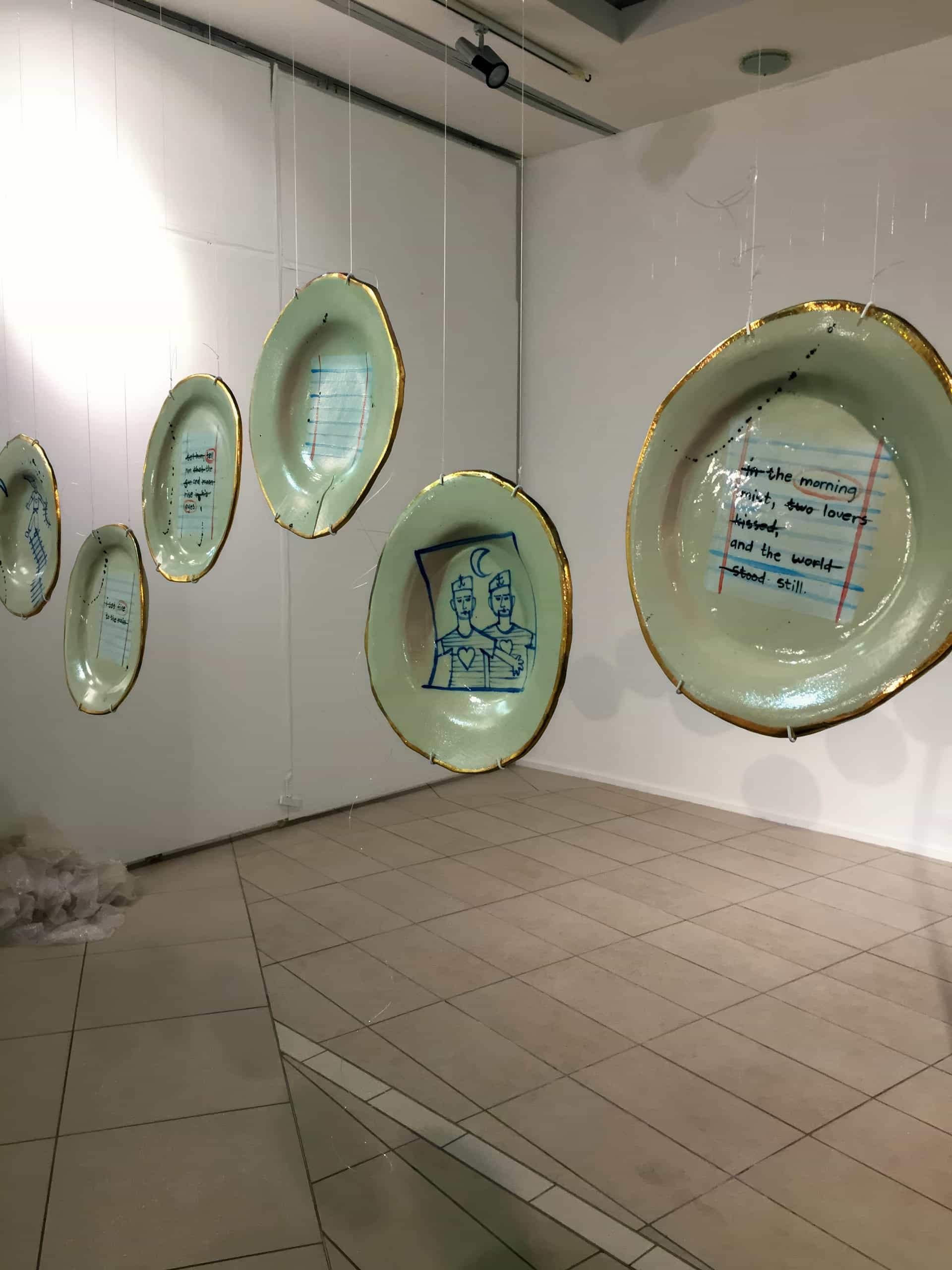 Several large, translucent plates hang in a row from the ceiling in an art gallery. Each plate has gold edges and features blue line drawings or handwritten text, including an illustration of two figures and poetic lines. The plates cast faint shadows on the walls and floor.