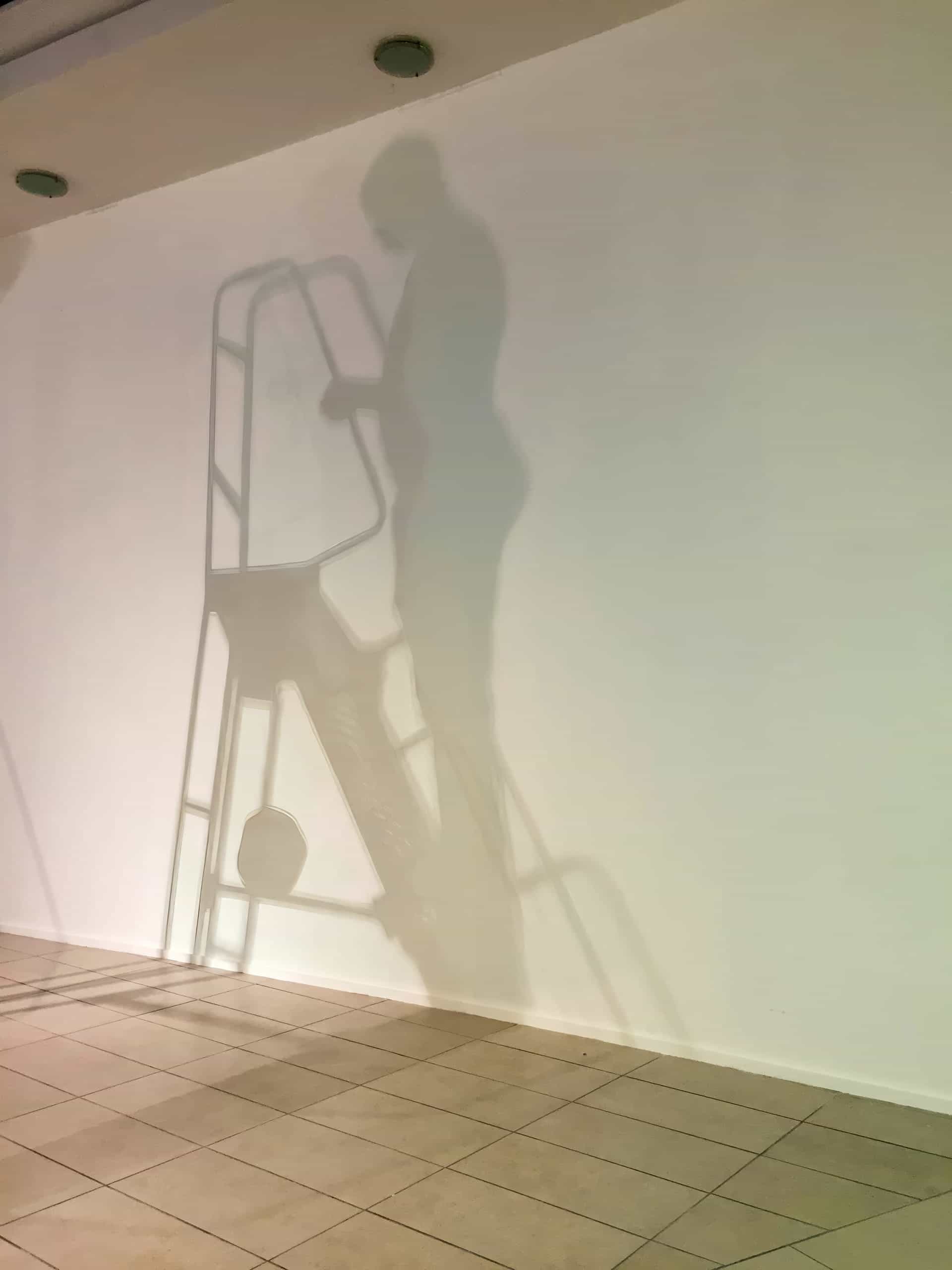 A shadow of a person standing on a step-ladder is cast on a blank white wall. The tiled floor and ceiling lights are also visible, but the actual person and ladder are not shown—only their clear silhouettes.