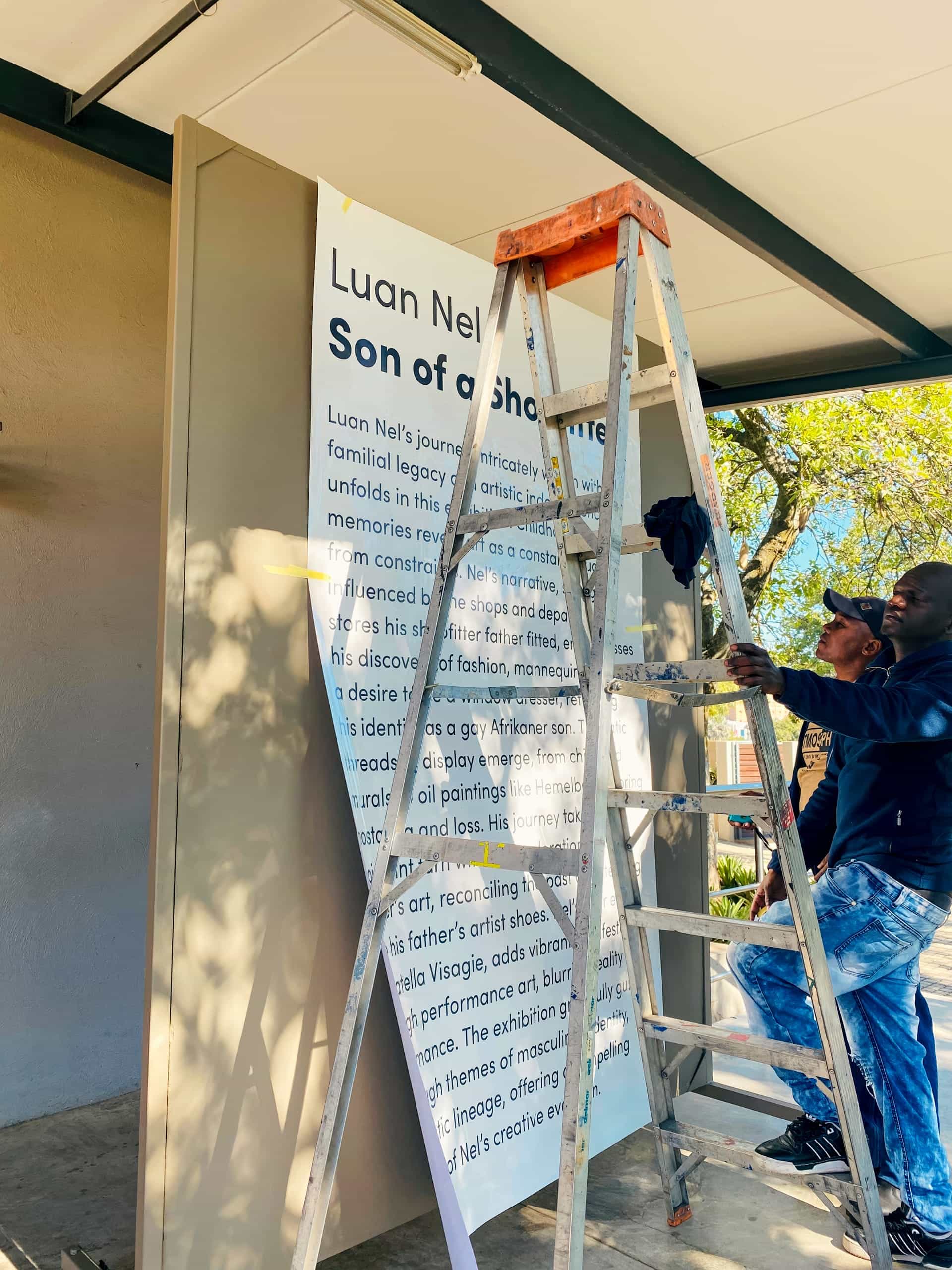 Two men stand beside a metal ladder, installing or examining a large signboard on a building’s exterior. The partially visible sign reads “Luan Nel... Son of a Sho...” and features text about an artistic journey. Sunlight casts shadows, and trees are in the background.
