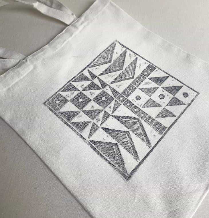 A white fabric tote bag lies flat on a neutral surface. The bag features a large, square geometric design in black ink by Contra.Joburg, displaying patterns of triangles, dots, and lines for an abstract Weight of Silence effect.