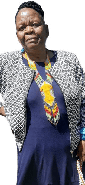 A middle-aged woman stands facing the camera, wearing a navy blue dress and a patterned jacket. She has short hair and a colourful, geometric beaded necklace. Her calm expression reflects the Weight of Silence against a plain, light background.