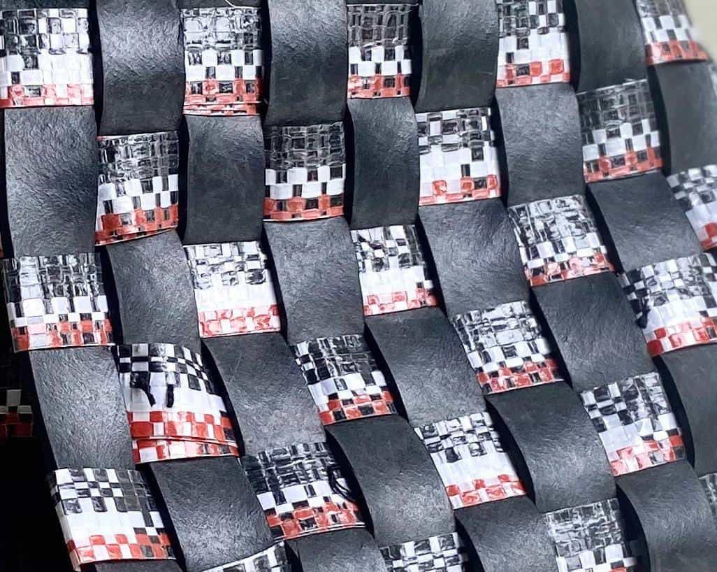 A close-up of an interwoven pattern made of black, glossy strips and metallic tape with a chequerboard design in silver, white, black, and red—echoing the Weight of Silence in its tight, basket-like structure with a slightly curved surface.