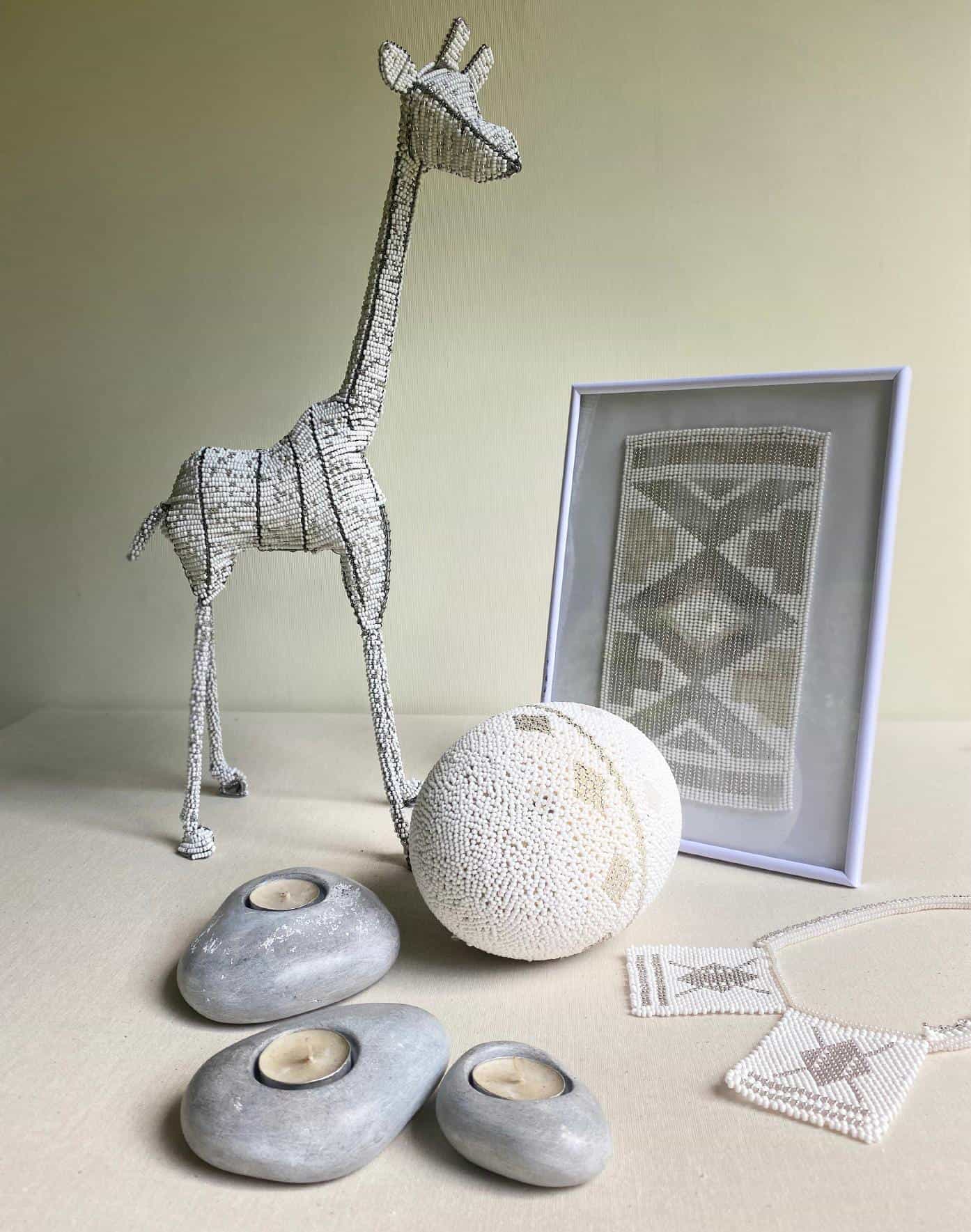 A beaded giraffe sculpture, beaded egg, and framed beadwork from Contra.Joburg sit on a light table. Nearby, smooth grey stone candle holders and a beaded necklace evoke the Weight of Silence in this minimalist, neutral-toned display.