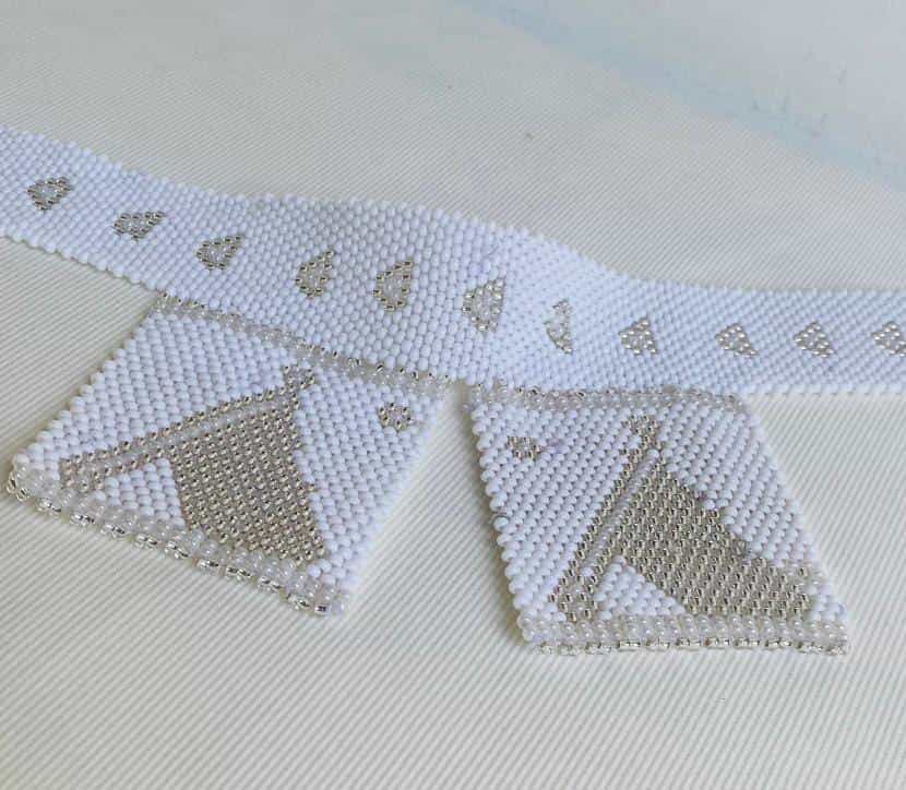 A white beaded belt from the Weight of Silence collection features two pendant-like rectangular sections. Delicate silver and white bead patterns, seen at Contra.Joburg, showcase crescents and abstract designs on a textured white background.