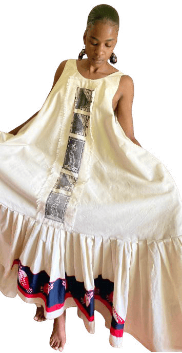 A person wearing a long, sleeveless white dress with a flared bottom and ruffled hem from Contra.Joburg. The dress features vertical decorative panels and a wide navy, red, and white patterned band near the hem. The person is barefoot—a study in the Weight of Silence.