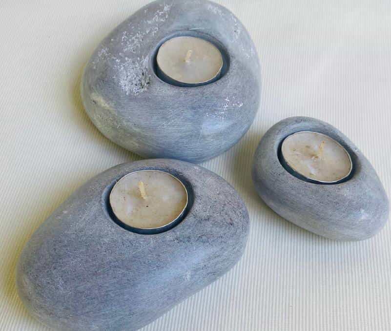 Three smooth, grey stones from Weight of Silence, each with a round cavity holding an unlit tealight candle, are arranged on a light-coloured, textured surface. The stones vary in size and have a natural, matt finish.
