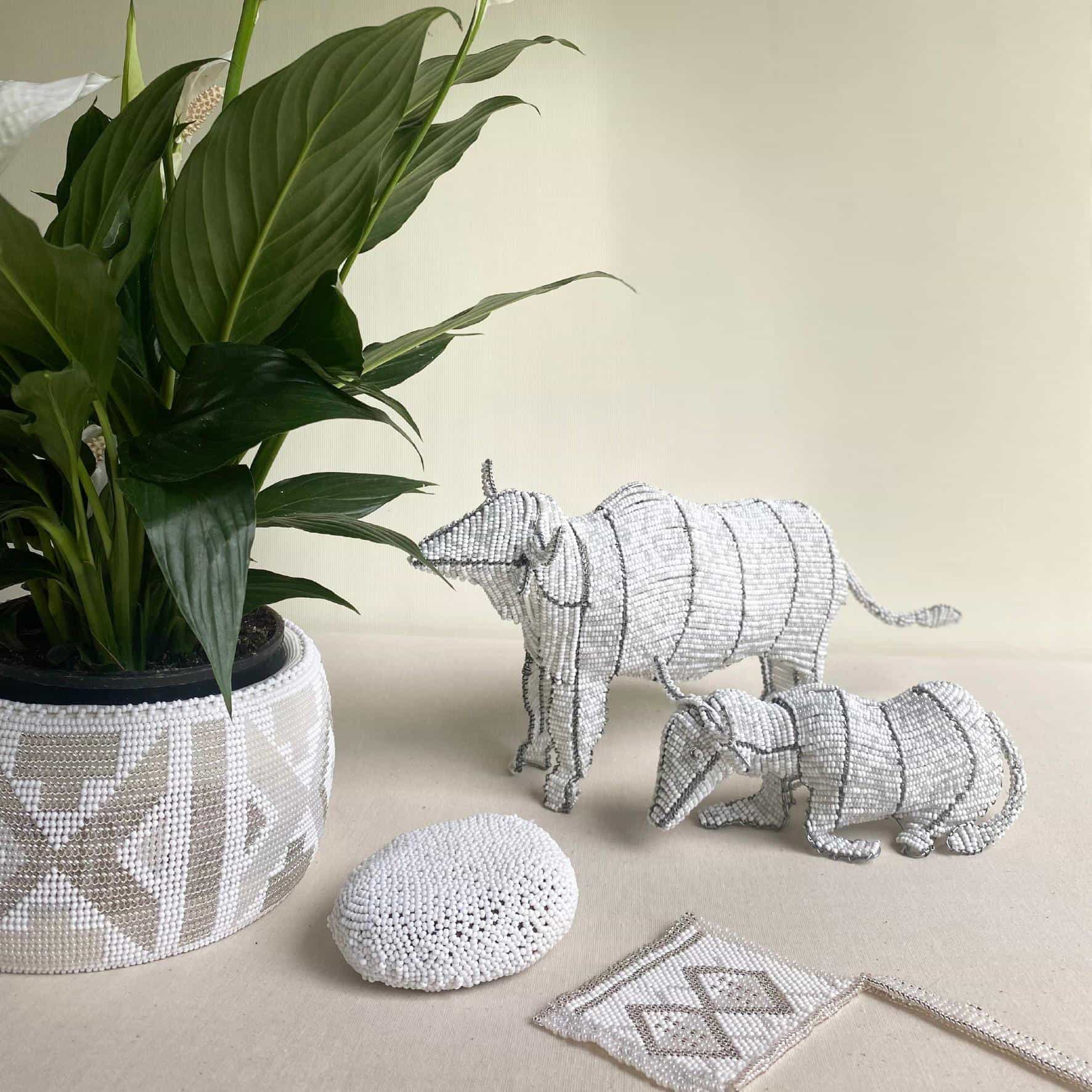 A potted plant with broad green leaves sits in a beaded white and grey pot cover. Beside it, two beaded animal sculptures from Contra.Joburg—a cattle adult and calf—stand near a beaded stone and a pouch patterned like Weight of Silence.