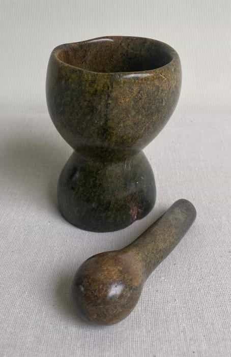 A stone mortar and pestle set sits on a light fabric surface, its smooth, greenish-brown finish evoking the calm Weight of Silence—a fitting accent for any Contra.Joburg-inspired kitchen.