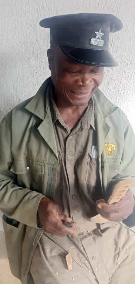 An older man in a uniform with a cap and jacket, smiling whilst whittling wood. Against a light textured wall, wood shavings cover his lap. The star insignia hints at memories—The Weight of Silence quietly present on his embroidered jacket.