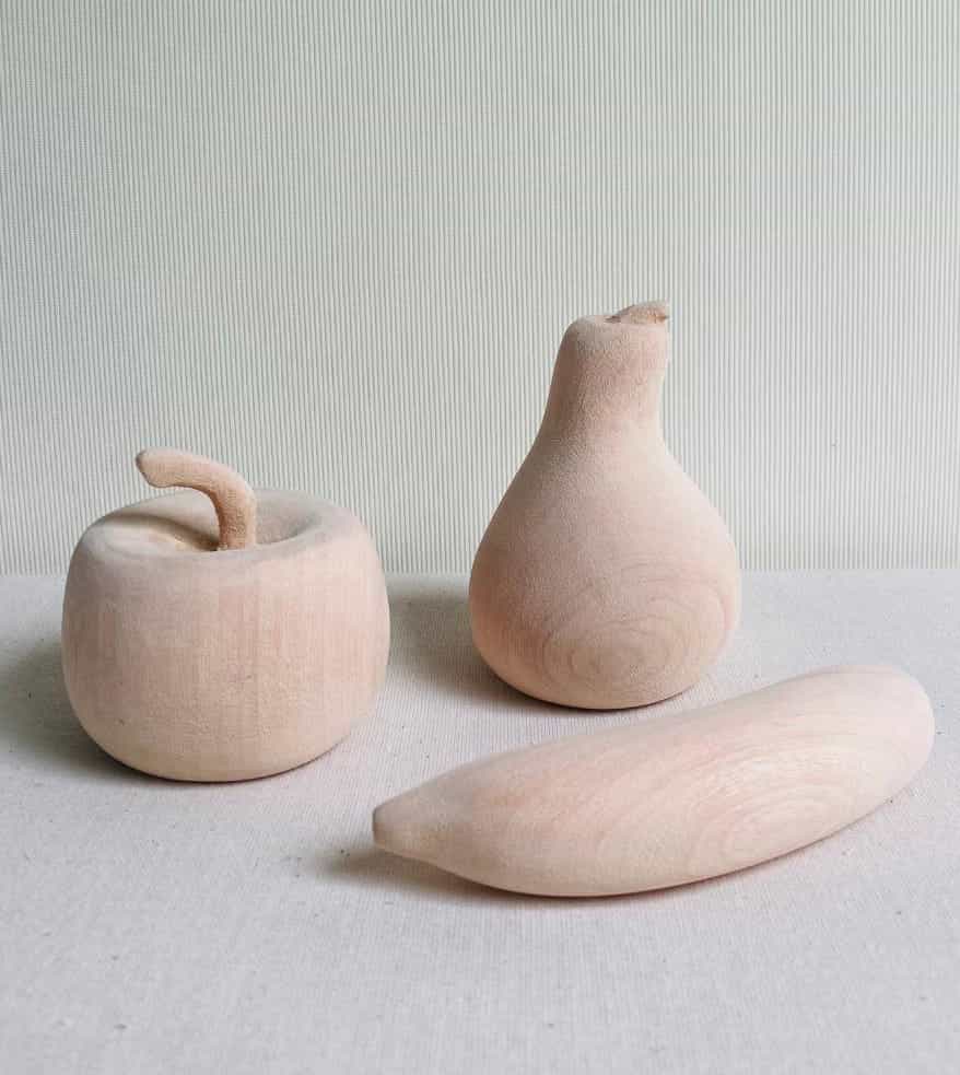 Three wooden sculptures shaped like fruit—a round apple with a curved stalk, a pear standing upright, and a banana lying on its side—sit quietly like the Weight of Silence on a light textured surface with subtle stripes, their smooth pale wood revealing graceful grain.