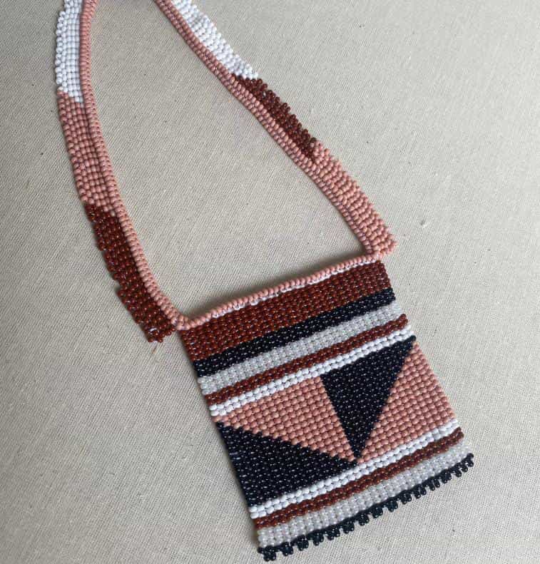 A beaded necklace from Contra.Joburg with a rectangular pouch, featuring geometric patterns in dusty pink, white, black, and dark brown beads. Named Weight of Silence, it’s displayed on a cream fabric background.