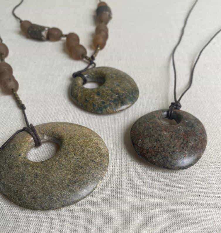 Three stone pendant necklaces lay on a light fabric surface, their earthy tones evoking the Weight of Silence. Each smooth, round stone with a central hole is strung on dark cord—one accented by irregular brown beads—crafted in the spirit of Contra.Joburg.