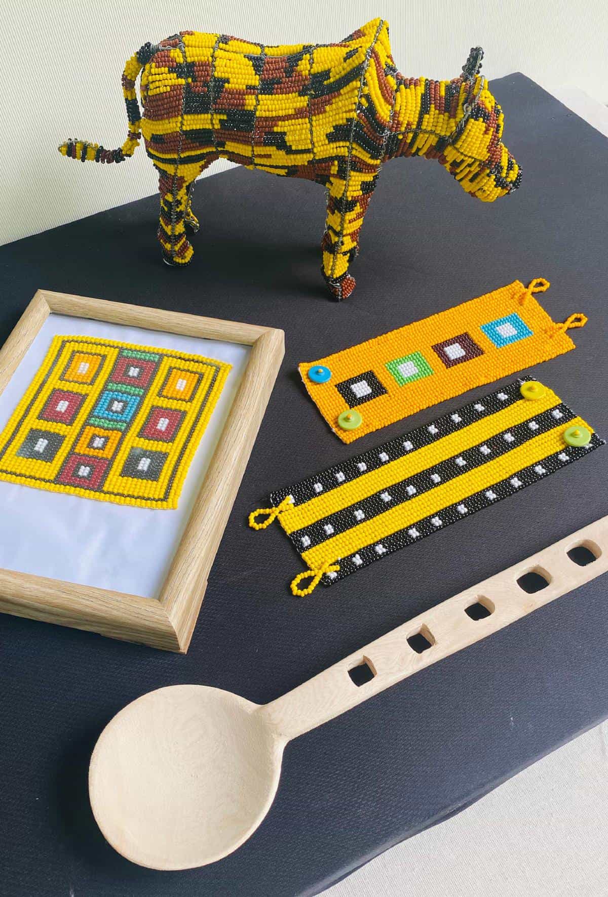 A beaded cow sculpture, a framed geometric beadwork design, three colourful beaded bracelets, and a wooden spoon with square cut-outs are displayed on a black surface for Contra.Joburg’s Weight of Silence, featuring bright patterns in yellow, black, green, and blue.