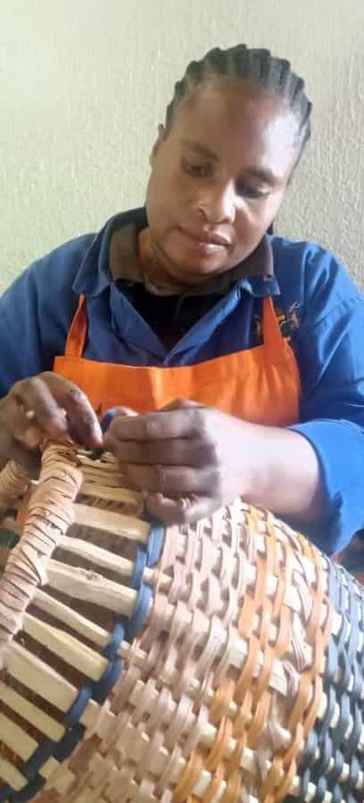 A woman wearing a blue shirt and orange apron, part of Contra.Joburg, focuses on weaving a basket from light and dark natural materials, her hands moving with the weight of silence as she intricately intertwines the fibres in a softly lit setting.