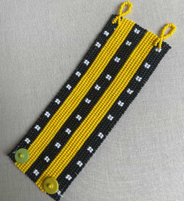 A beaded bracelet featuring alternating black and yellow vertical stripes, inspired by Contra.Joburg. The black stripes have small white bead clusters, whilst solid yellow beads and green buttons evoke the subtle Weight of Silence in its design.
