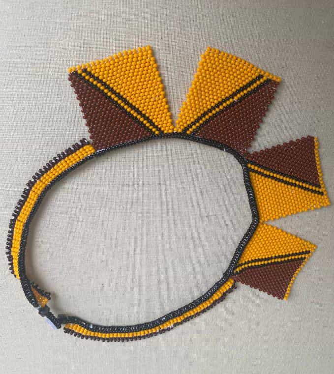 A beaded necklace with triangular fringe pieces in yellow and brown. The striking design, reminiscent of the Weight of Silence collection by Contra.Joburg, features alternating triangles on a neutral fabric background for a bold statement look.