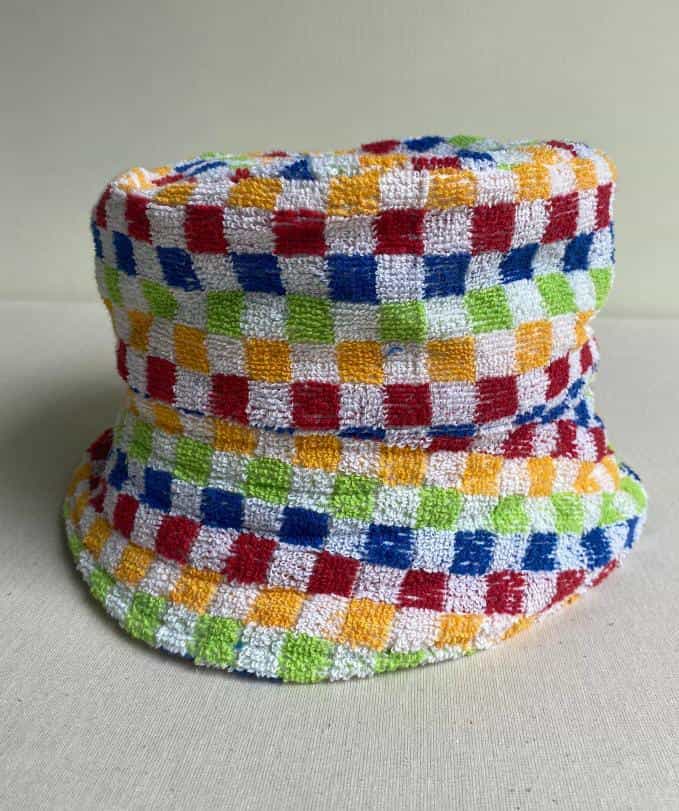 A bucket hat made of textured fabric featuring a colourful chequerboard pattern with alternating squares of red, blue, yellow, and green on a white background—an eye-catching piece inspired by the playful spirit of Contra.Joburg.