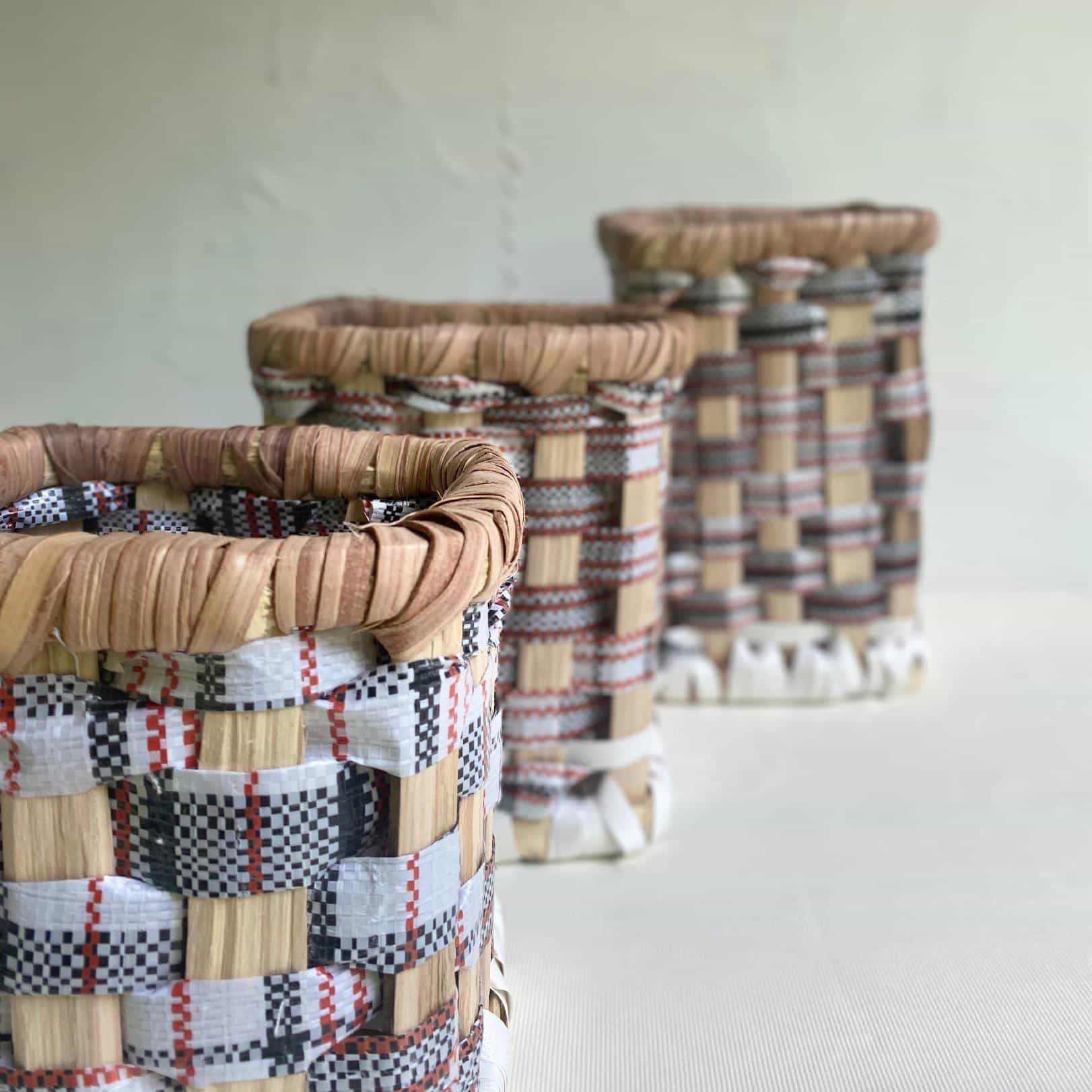Three woven baskets with chequered patterns in natural, white, red, and black tones are arranged in a diagonal line on a light surface. Their textured, handwoven details echo the artistry of Contra.Joburg. The background is softly blurred.