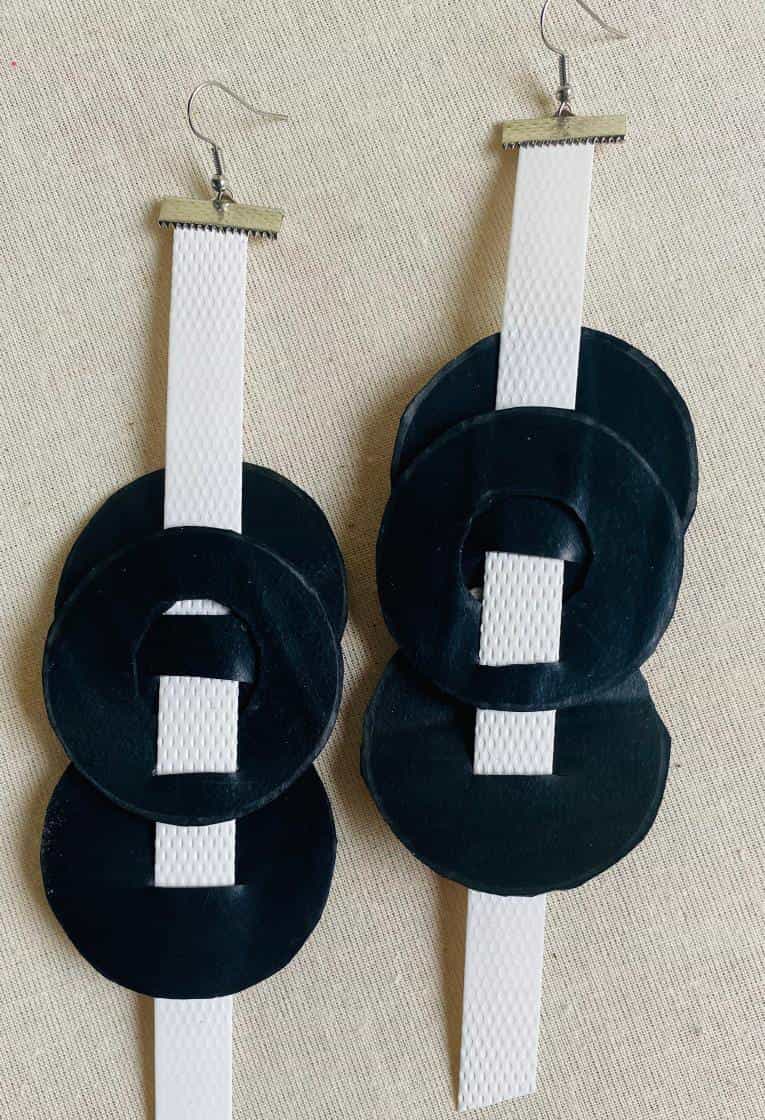A pair of handmade earrings from Contra.Joburg features white textured vertical strips with three overlapping black velvet circles on each, capturing the Weight of Silence. The earrings have silver hooks and are displayed on a beige fabric background.