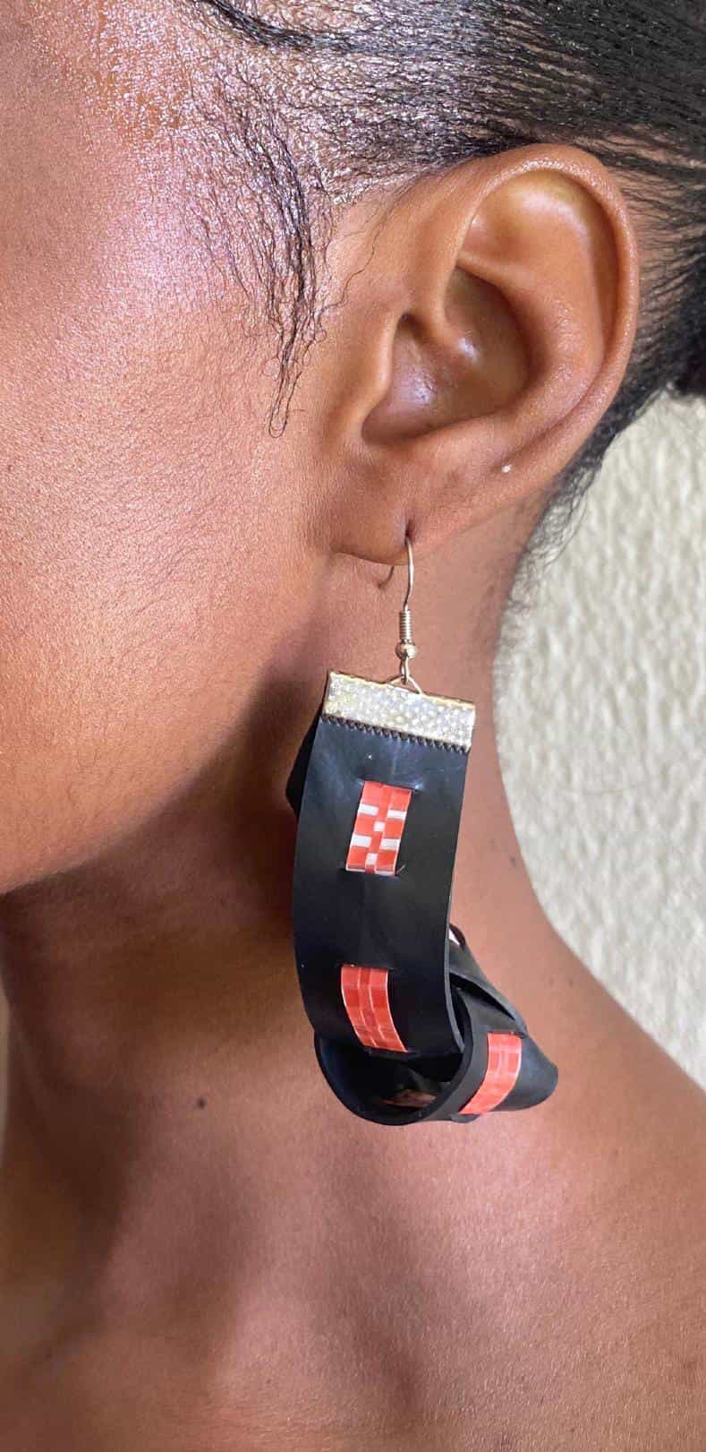 A close-up of a person's ear wearing a large, handmade black earring with red square designs and a metallic hook. Crafted from upcycled material, this Contra.Joburg piece loops in an artistic shape. The person has short hair.