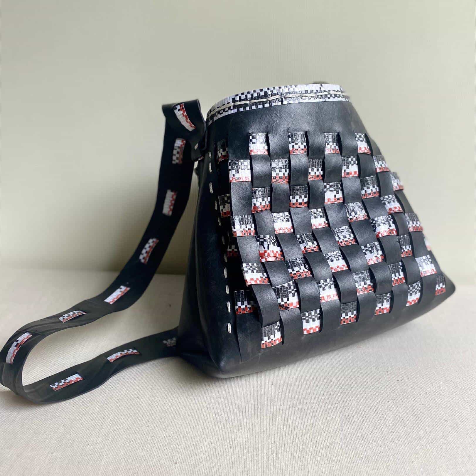 A stylish shoulder bag made of black leather with a basket-weave pattern, featuring interlaced strips of black, white, and red material. The adjustable Contra.Joburg strap displays a matching patterned design. The bag is placed against a light beige background.