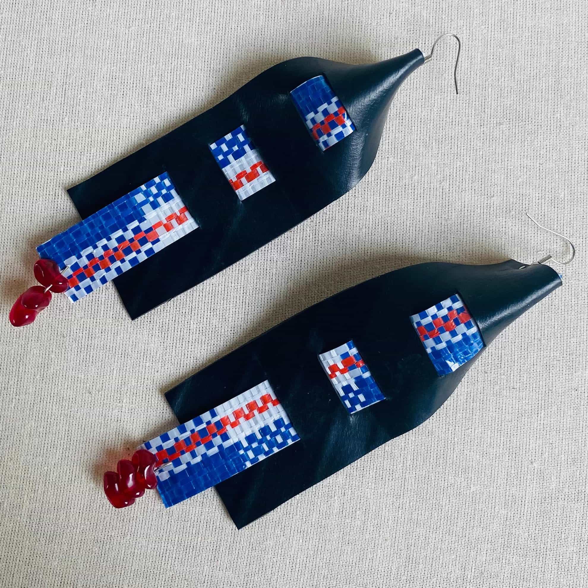 Two long, black earrings lay on a light textured surface. Their blue, white, and red beaded designs evoke the modern, geometric style seen at Contra.Joburg, whilst small pink beads and metal hooks complete this striking Weight of Silence-inspired look.