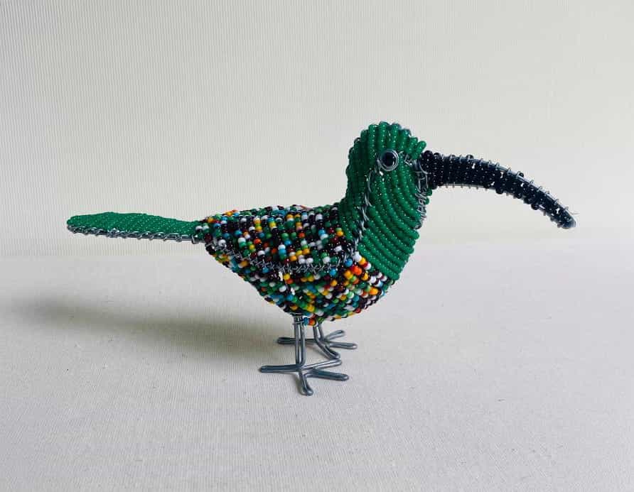 A beaded bird sculpture from Contra.Joburg, with a long, curved black beak, green head and tail, and a multicoloured body stands on wire legs against a plain background—evoking the Weight of Silence in its intricate beadwork.