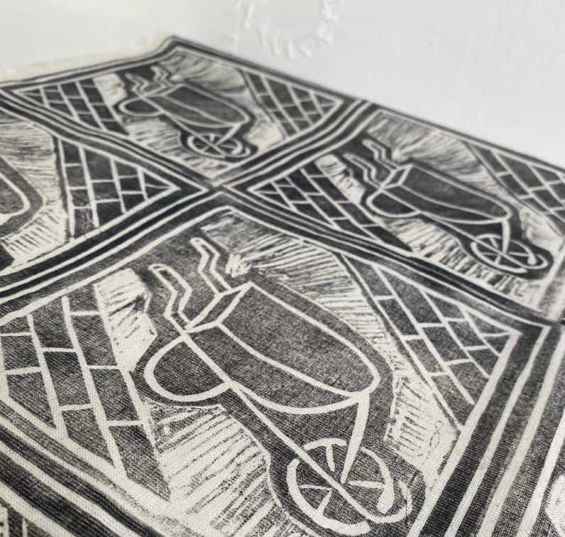 Black-and-white fabric from Contra.Joburg features repeated block-print wheelbarrow designs viewed from above, with geometric patterns in the background. Draped near a white textured wall, the image highlights close-up details of this Weight of Silence print.