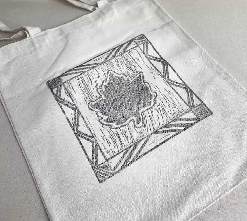 A white canvas tote bag lies flat. The bag features a black, hand-printed design of a maple leaf in the centre, framed by a square border with patterns inspired by Weight of Silence and geometric shapes from Contra.Joburg.
