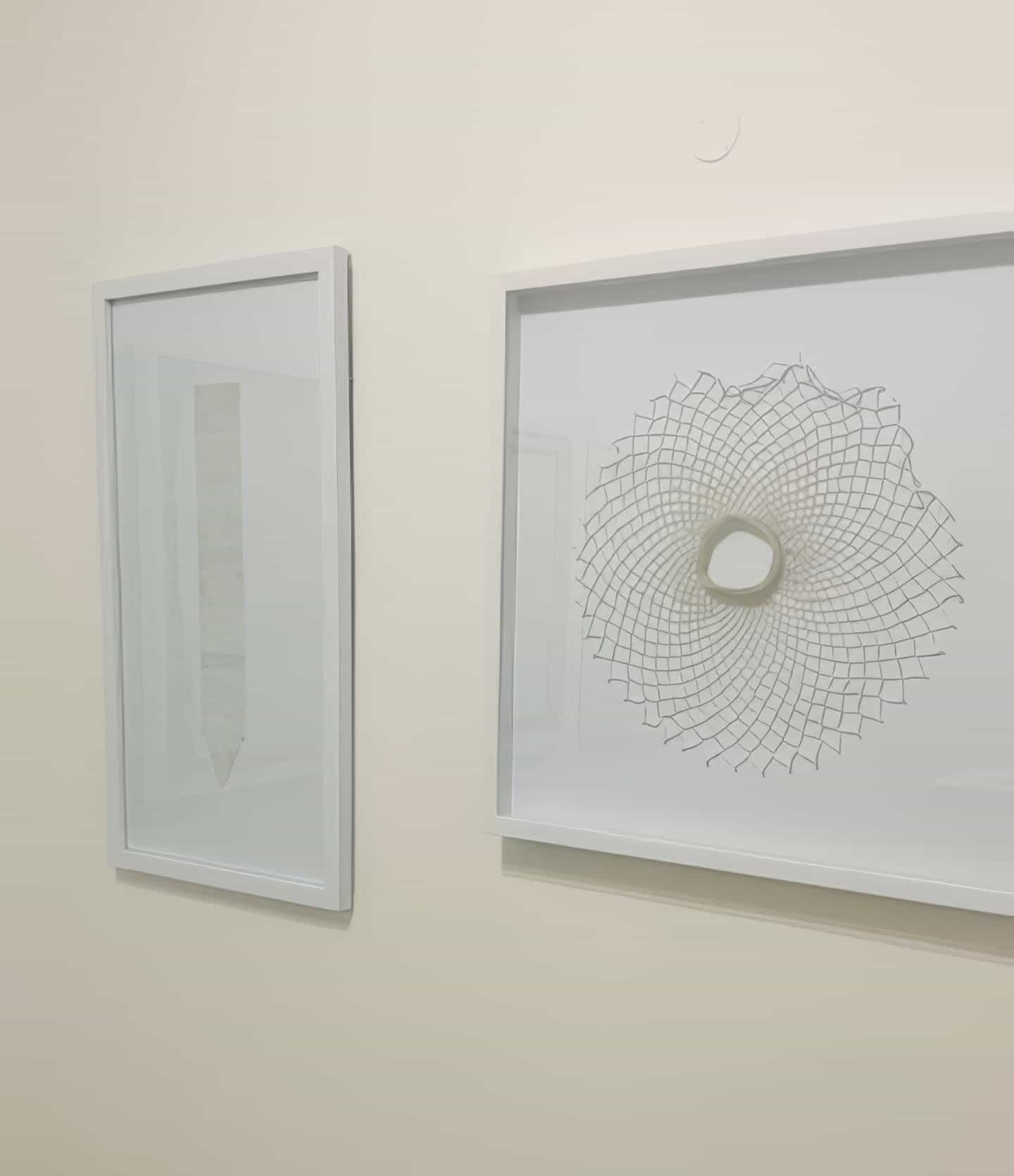 Two framed abstract artworks hang on a pale wall at Contra.Joburg. The left piece features a vertical, pencil-like shape—echoing the Weight of Silence. The right displays a spirograph pattern with an irregular central opening; both frames are white.