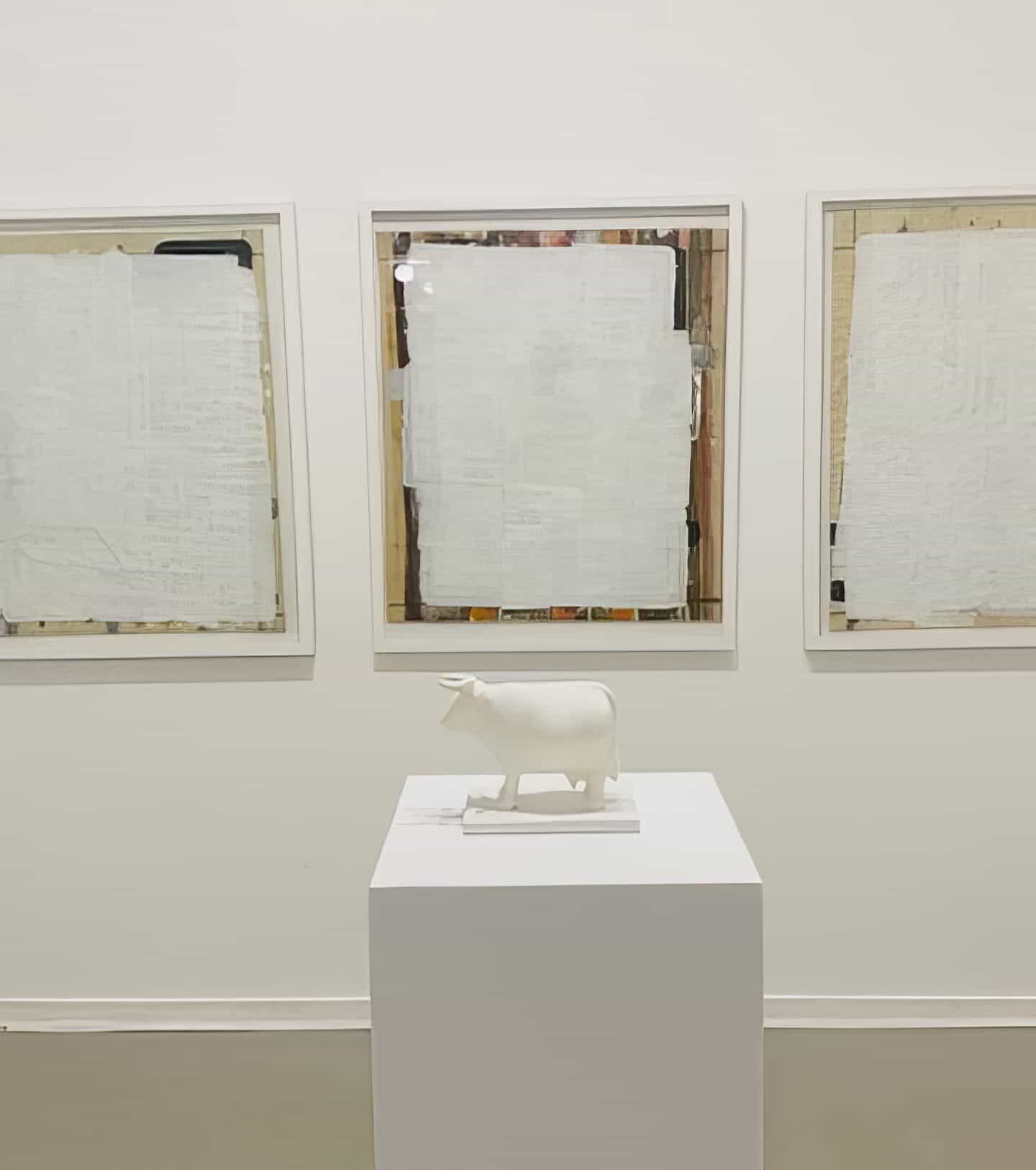 A small white sculpture of a cow, part of Contra.Joburg’s Weight of Silence series, stands on a plinth before three framed artworks—each with textured, layered surfaces that veil hidden details—all set against a plain white wall.
