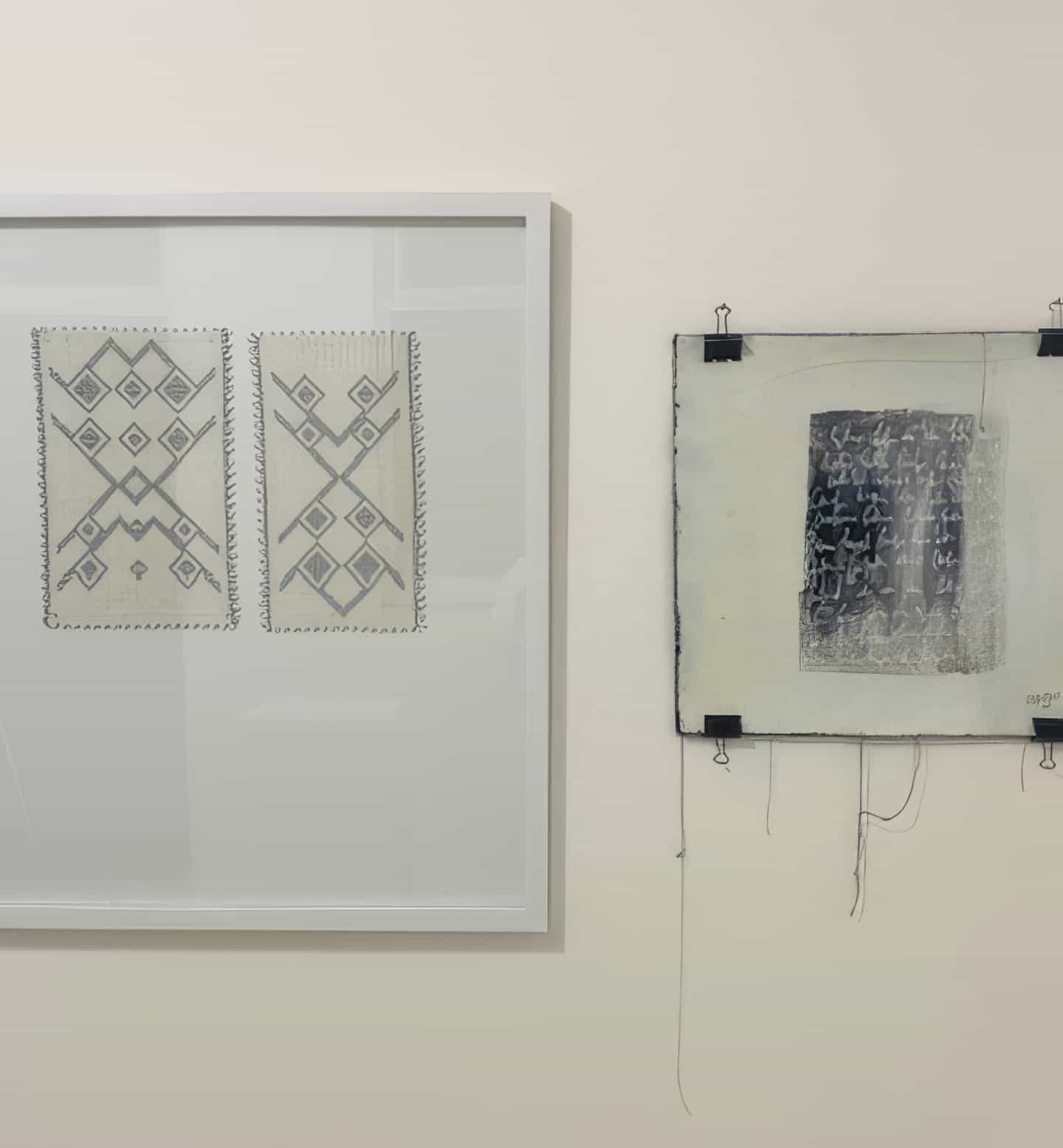 Two framed artworks hang on a white wall at Contra.Joburg. The left piece shows geometric, symmetrical patterns, whilst the right, titled Weight of Silence, features abstract dark markings with loose threads and black clips.
