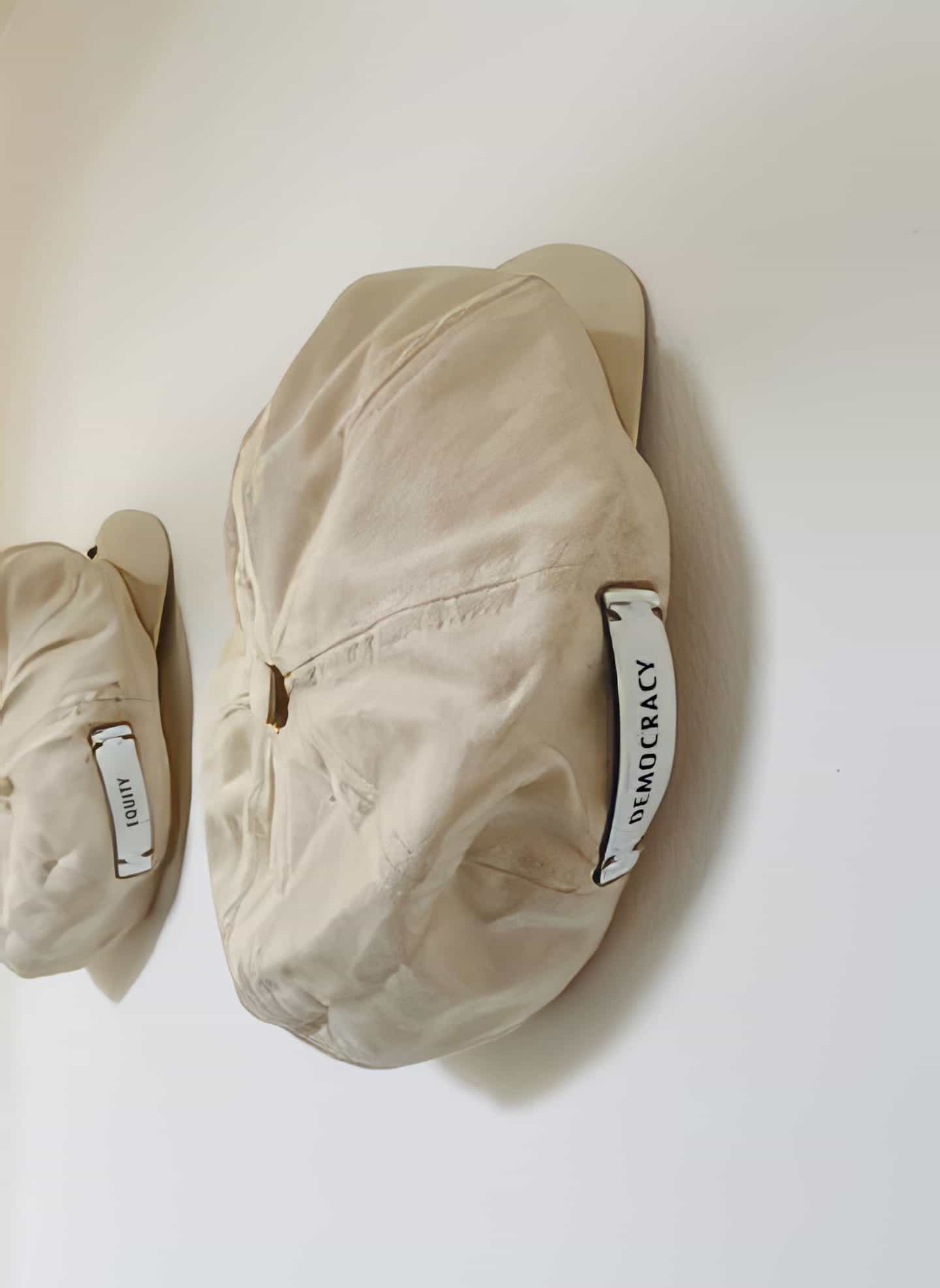 Two off-white, wrinkled caps hang on wooden wall pegs against a white background. A white label with “DEMOCRACY” in black text recalls Contra.Joburg’s Weight of Silence, while the farther cap’s label remains partially visible and unreadable.
