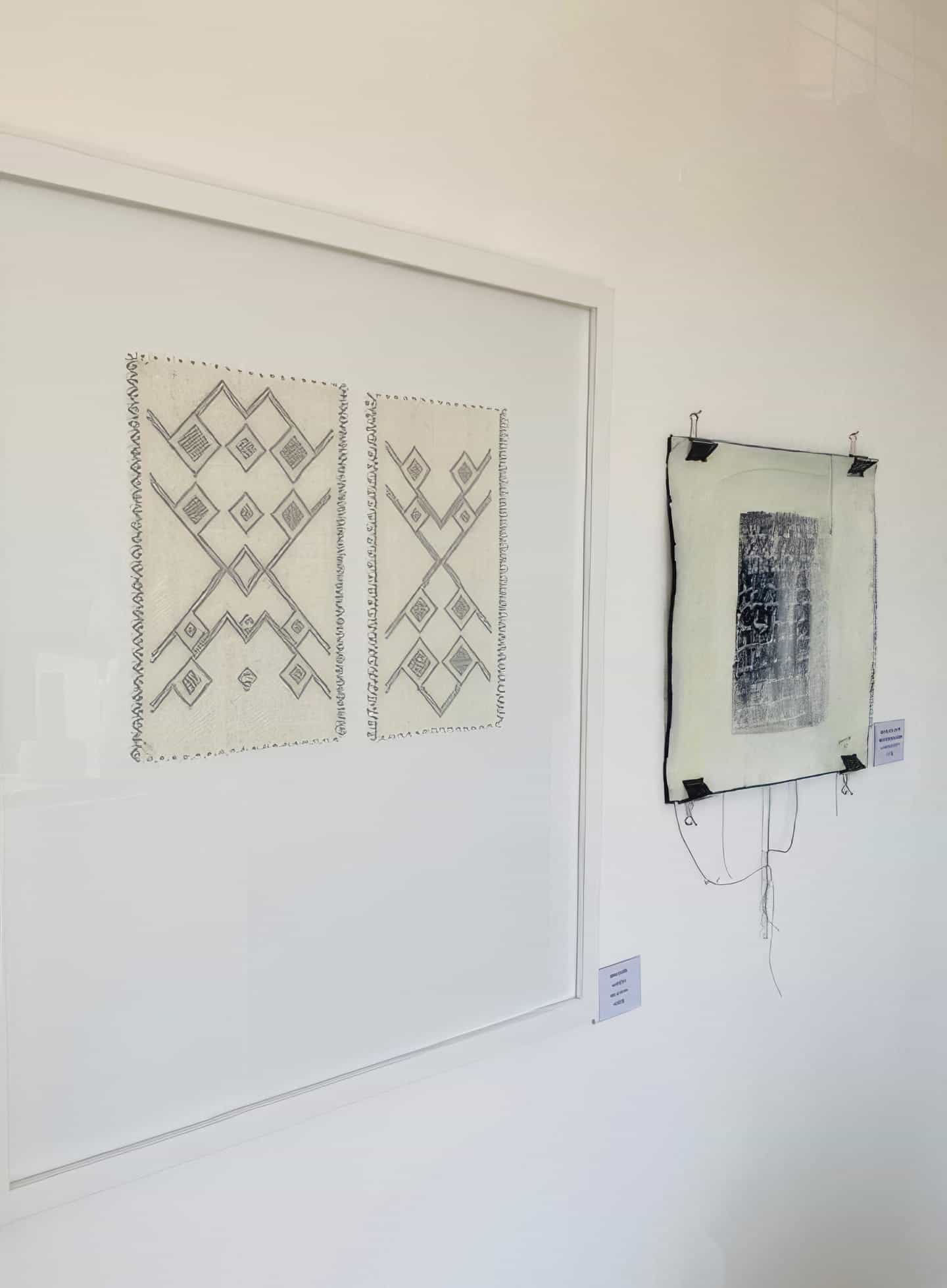 Two framed abstract artworks hang on a white wall. The left piece, Weight of Silence, features black ink diamonds and zigzags. The right artwork, part of Contra.Joburg, has a textured dark rectangle pinned to off-white fabric with loose threads.