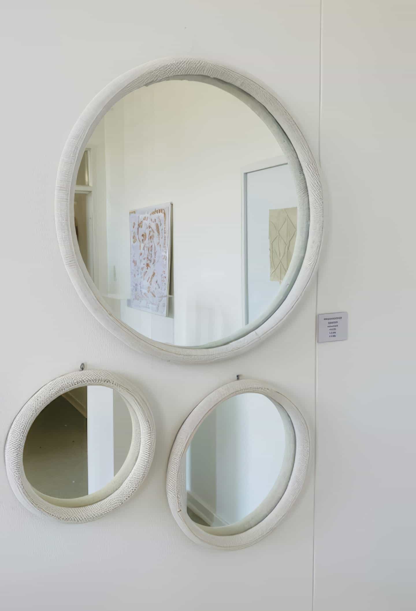 Three round mirrors with textured white frames are mounted on a white wall at Contra.Joburg. Reflections reveal a framed abstract artwork and a doorway, while a small wall label hints at an art display, possibly part of 