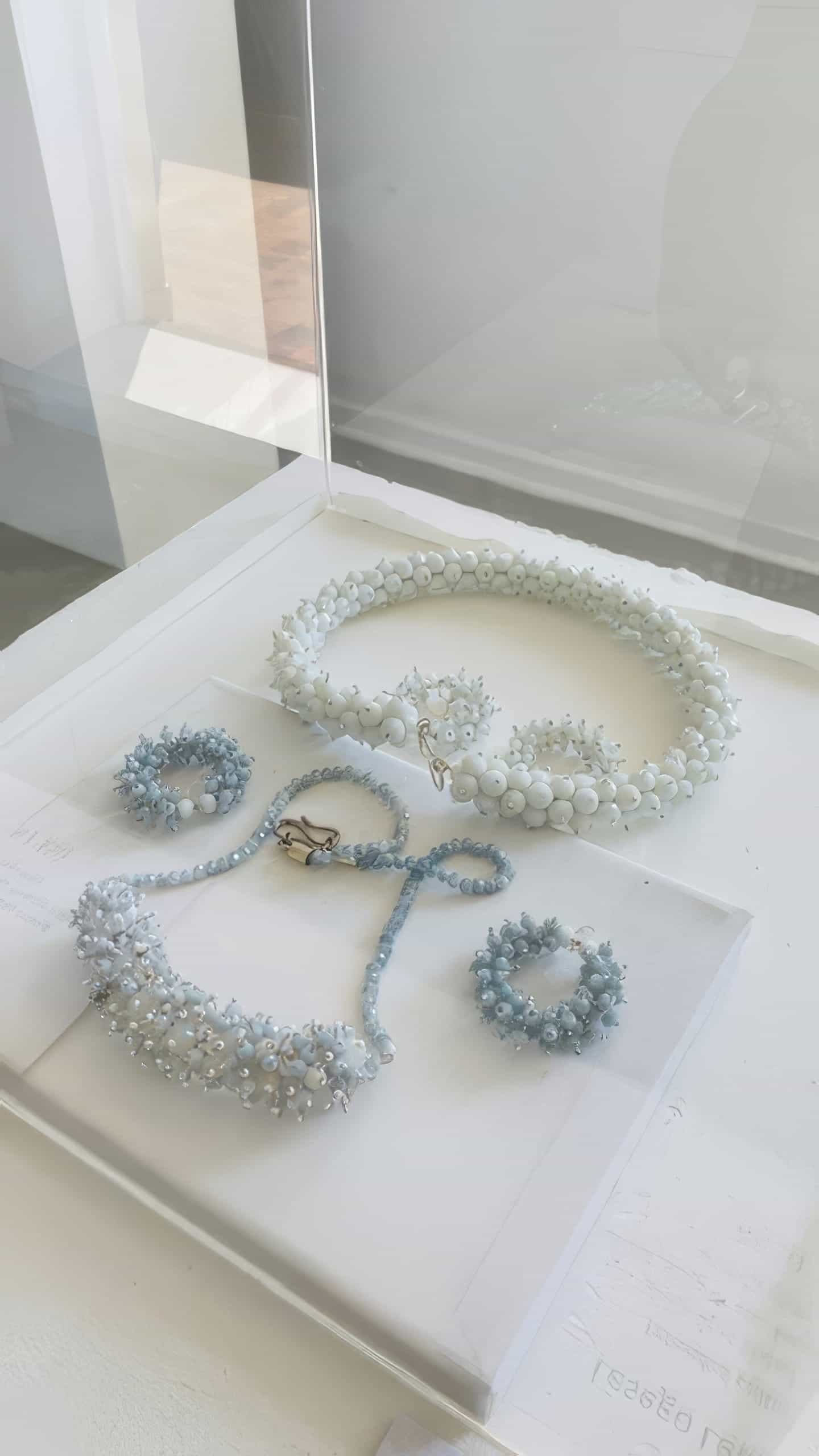 A jewellery display case with a necklace, bracelet, ring, and earrings from Contra.Joburg. Each piece features white and pale blue beads and pearls, creating a delicate floral texture. The clean display is set by a sunlit window, evoking the Weight of Silence.