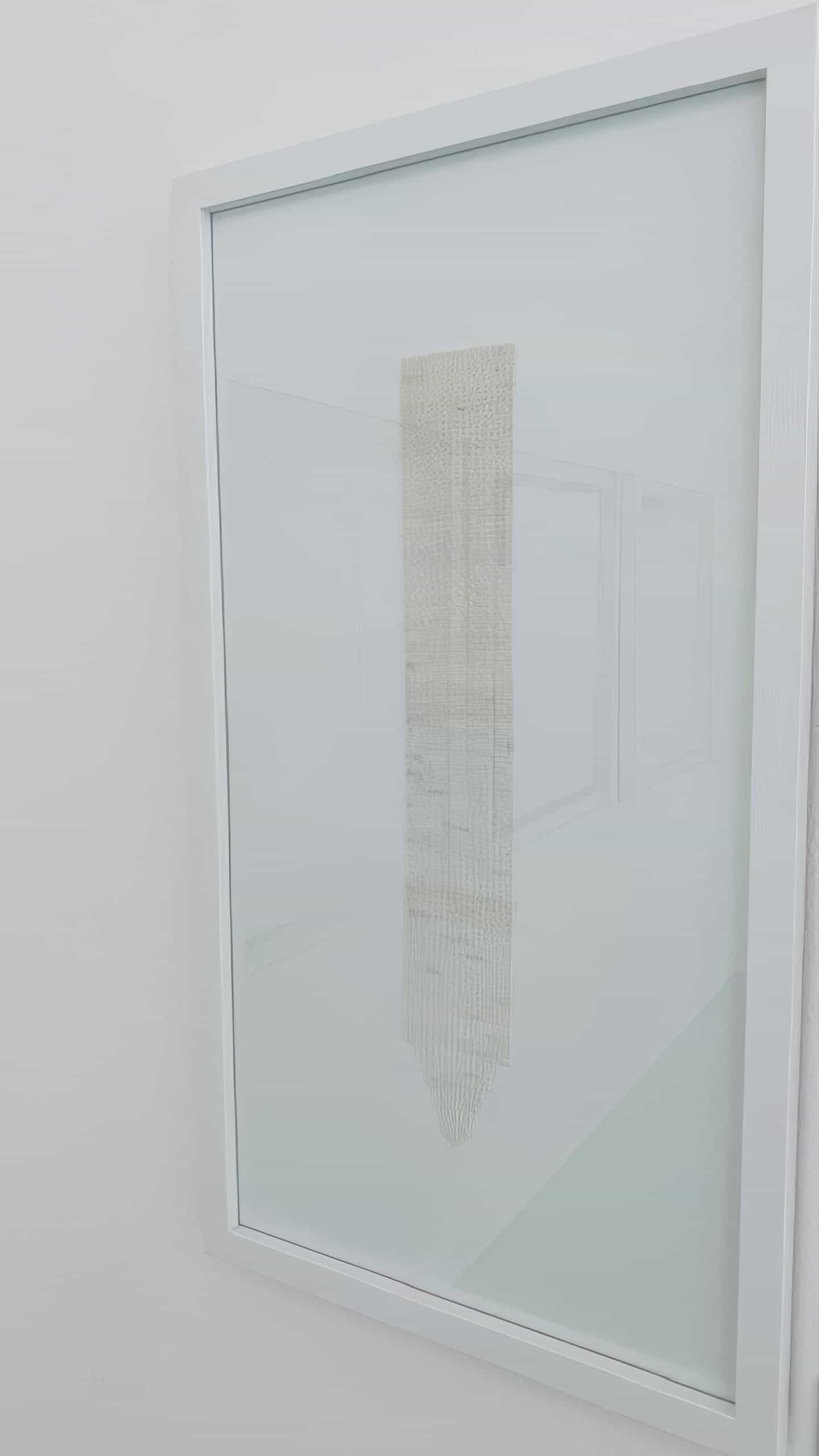 A white-framed glass case displays a narrow, vertically hung, textured textile piece in light beige tones. The minimalist background and faint reflections highlight the Weight of Silence, emphasising the simplicity and delicacy of the artwork.