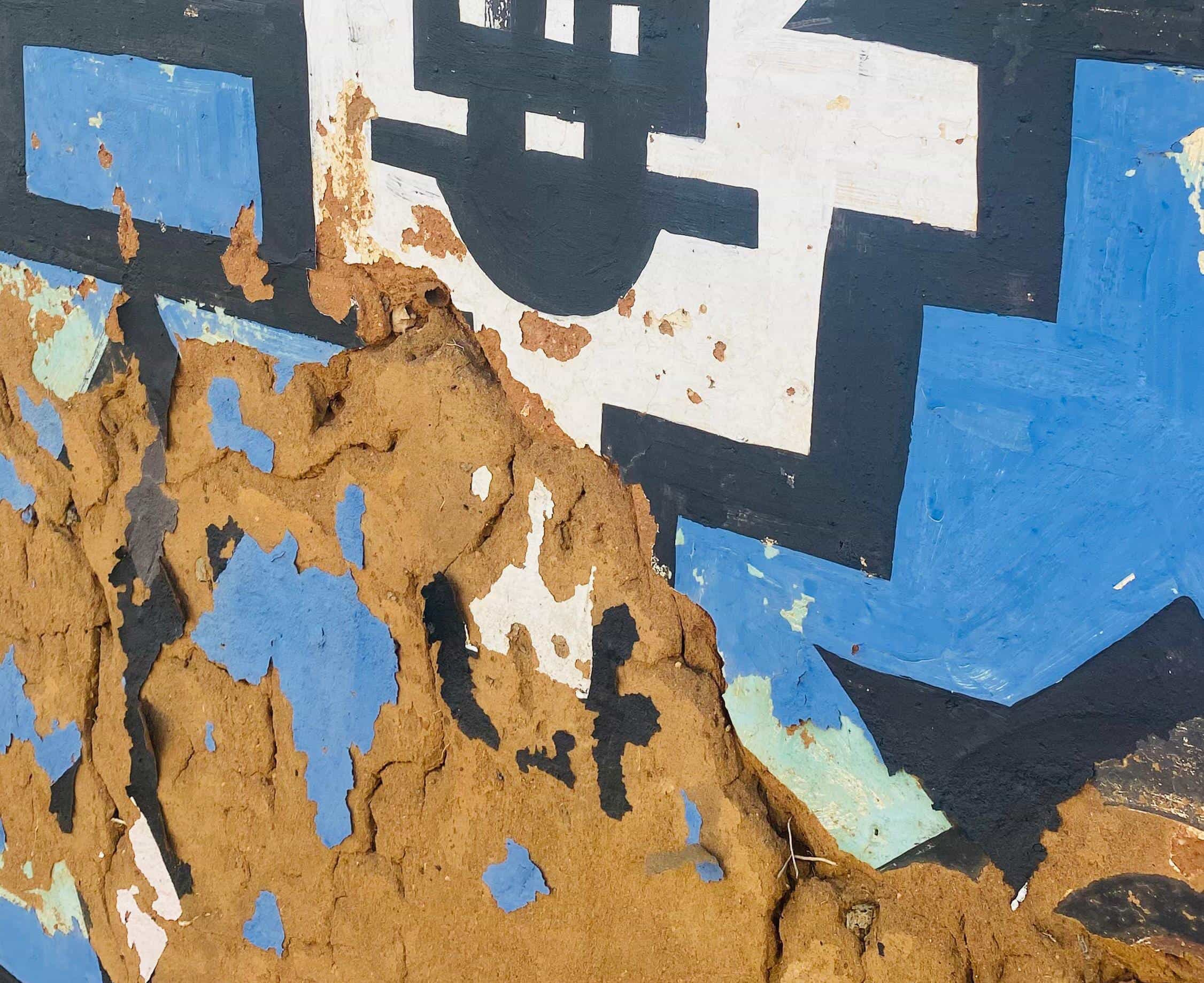 A close-up of a painted wall with geometric black, white, and blue patterns. The chipped paint reveals rough brown mud beneath, giving a weathered look—echoing the enduring spirit celebrated in the Legacy Project of Gogo Mahlangu.