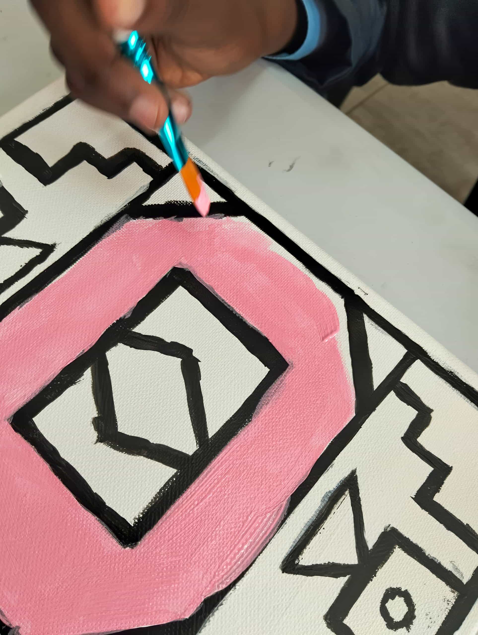 A hand holds a blue paintbrush, painting a large pink letter 