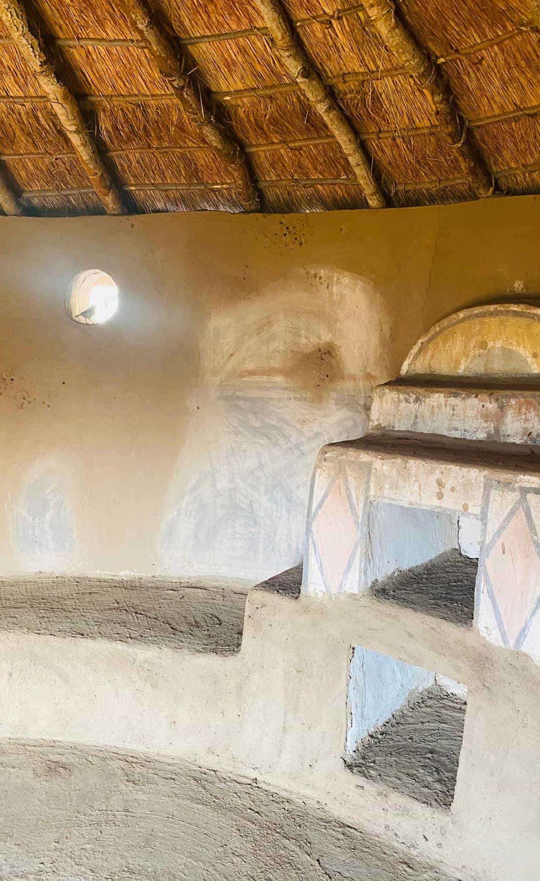 Interior of a traditional mud hut with a thatched roof, inspired by Gogo Mahlangu’s Legacy Project. Earth-toned walls with geometric patterns, natural light from a small window, and built-in mud benches create a rustic, warm atmosphere perfect for celebration.