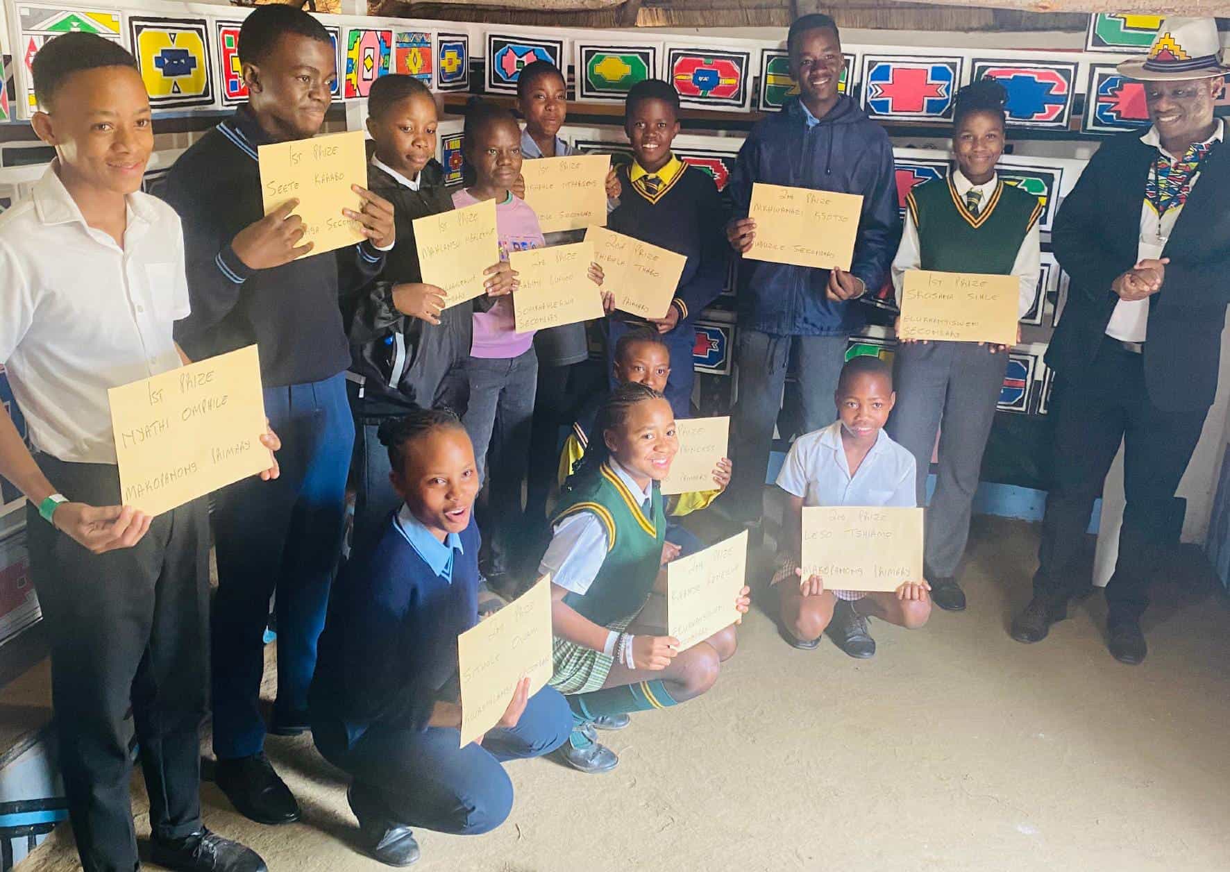 A group of students and two adults pose indoors, smiling and holding certificates during a Legacy Project celebration. The students wear school uniforms, the adults are in formal attire, and colourful artwork adds a vibrant backdrop to the scene.